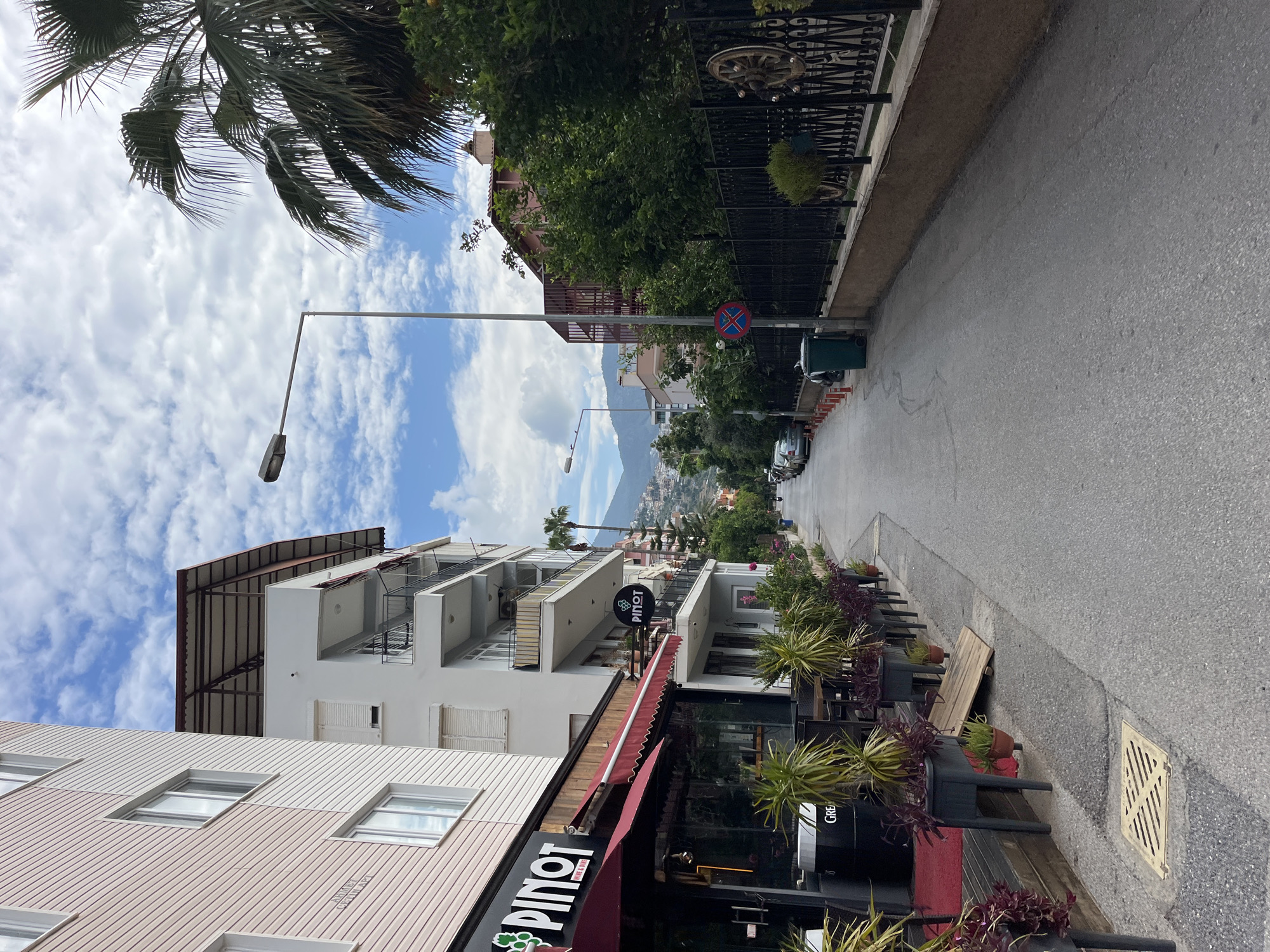 The image captures a street scene in Alanya, Turkey. The view is along a narrow, paved road heading uphill, with buildings on either side. On the left, there's a multi-story building with a restaurant named "Pinot" at street level, indicated by signage. The building has balconies and what appears to be exterior shutters. The right side of the road features a fence with decorative elements, trees, and shrubs. In the distance, a mountain range is visible beneath a partly cloudy sky. A street lamp is positioned on the left. Several cars are parked along the roadside. There's a pedestrian crossing sign and a small green trash bin on the right side of the street. Some plants are arranged in pots along the sidewalk next to the building on the right.