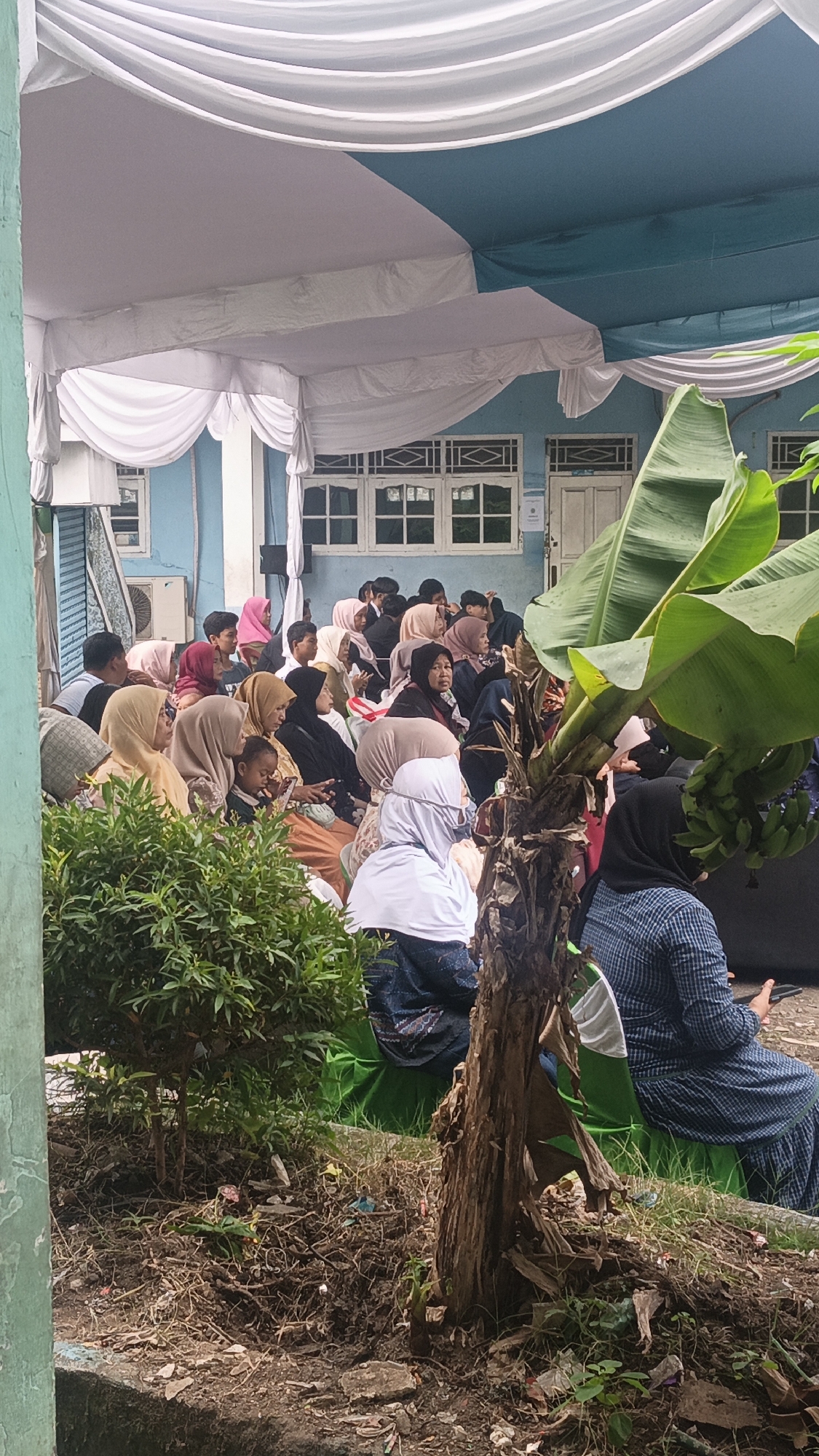 This image depicts an outdoor gathering of people, likely in Tigaraksa, Indonesia, possibly at a community event or religious ceremony. A group of individuals, predominantly women wearing hijabs and modest clothing, are seated on the ground, facing towards the left of the frame. They appear to be listening to something or observing an event, with some engaged in conversation. A child is visible among the seated individuals. The setting is under a temporary tent structure with white and blue fabric drapes. In the foreground, a small, leafy green plant and a thick, rough-barked tree trunk are prominent. The ground appears to be unpaved and scattered with debris, indicating an outdoor, potentially informal, environment. The background reveals a light blue building with windows and doors, suggesting it might be a community center or a school. The overall atmosphere appears to be one of communal gathering and shared activity.