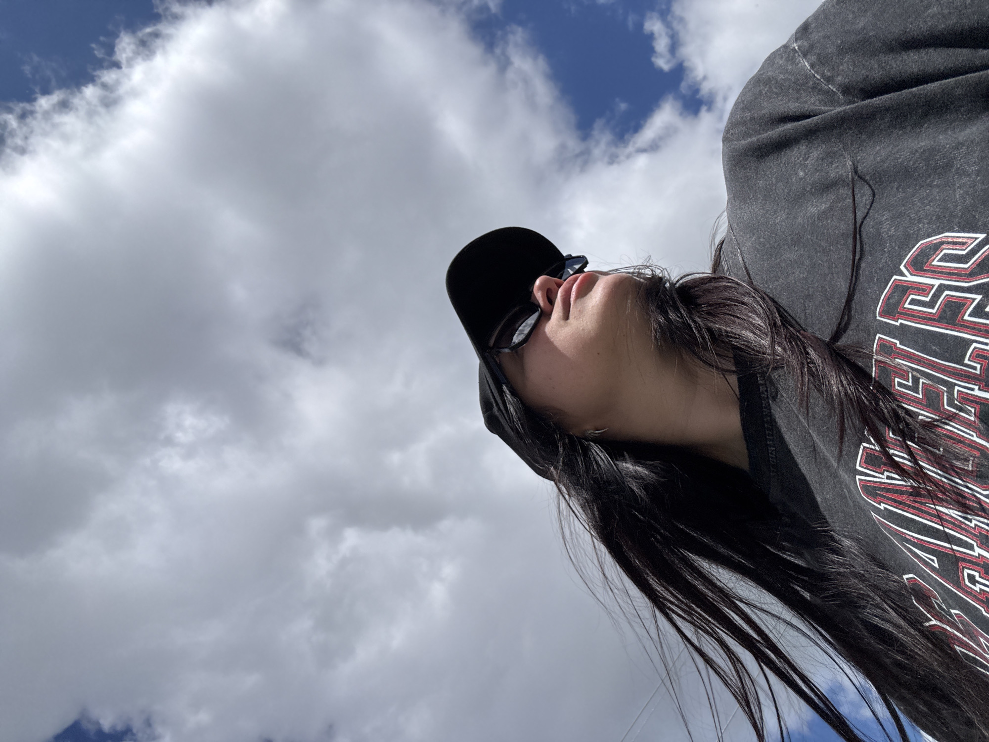 A low-angle photograph captures a person looking upwards against a backdrop of a partly cloudy sky. The subject, positioned in the upper right quadrant of the image, is seen from the chin up, wearing a dark baseball cap and dark sunglasses. Their head is tilted significantly backward, and their mouth is slightly ajar. Long, dark hair cascades down their back and shoulders.
The person is wearing a dark grey t-shirt with a distressed appearance. On the right side of the shirt, a prominent graphic features stylized red letters with white outlines. Although partially obscured by the person's hair and the fabric's folds, the letters "LLEGE" are clearly visible, strongly suggesting the word "COLLEGE" or a similar academic/athletic text.
The scene is set outdoors during the daytime under a bright sky. Large, white cumulus clouds are scattered across the blue sky, with visible patches of clear blue, especially towards the upper left. The bright lighting indicates a sunny day with some cloud cover. The perspective is from below, looking up, which gives a sense of openness and highlights the vastness of the sky. No specific location cues are visible beyond the sky and clouds.
