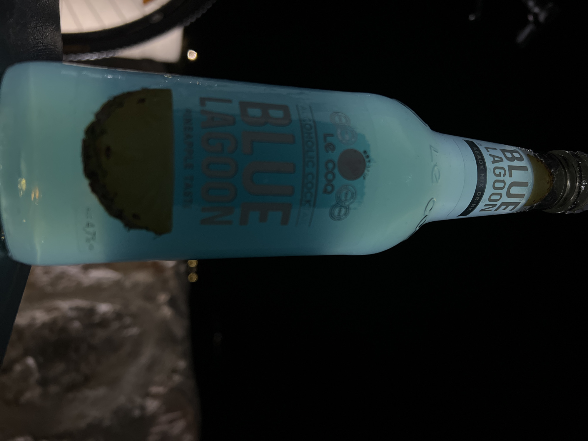 A horizontal shot depicts a bottle of "BLUE LAGOON" alcoholic cocktail drink. The bottle is light blue and features pineapple imagery. Text on the label includes "BLUE LAGOON", "LE COQ", "ALCOHOLIC COCKTAIL", and "READY MIX DRINK". The bottle is positioned at an angle, with its cap facing towards the right. The background is dark and blurry, with some hints of ambient light. There is no specific evidence to suggest the location is indeed Boreti, Montenegro; location is unknown.