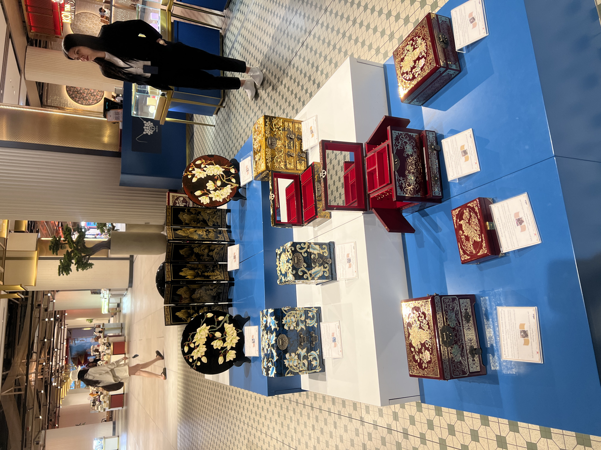 This image captures an indoor scene, likely a high-end shopping mall or exhibition space in Dubai, United Arab Emirates. The setting is bright and spacious, with a patterned tile floor in the foreground and polished light-colored flooring further back, leading to other commercial establishments.
The primary focus is an elaborate display of intricately decorated boxes and round side tables, showcased on white and blue platforms. These items appear to be traditional handicrafts, possibly lacquered wood or mother-of-pearl inlay pieces, suggesting an East Asian influence. The boxes feature rich colors such as deep red, gold, and dark blue, adorned with detailed designs including floral motifs, mythical creatures like dragons, and ornate scrollwork. Several boxes are open, revealing red velvet interiors with compartments, indicating they are likely jewelry or storage boxes. Two prominent round tables with black tops are embellished with delicate white magnolia flower designs. A multi-panel folding screen with gold and black artwork is partially visible behind one of the tables.
Next to the displayed items are several white information cards. One card clearly states "MOTHER-OF-PEARL PALACE AND GOOD FORTUNE BOX" and describes the item as a "traditional handicraft" with "symbols of good fortune." Another card partially shows "JEWELRY BOX." Small logos, possibly featuring flags or crests, are also visible on these cards.
Two women are visible in the scene. In the foreground on the left, a woman with long dark hair, dressed in a black suit jacket, white top, black trousers, and white shoes, is observing the display. Further back and to the left, another woman wearing a light-colored outfit and a white cap walks away from the display area. Other indistinct figures can be seen in the distant background. The activity suggests browsing or viewing an exhibition of these decorative items. The lighting is bright and artificial, typical of an indoor commercial environment during what appears to be daytime.