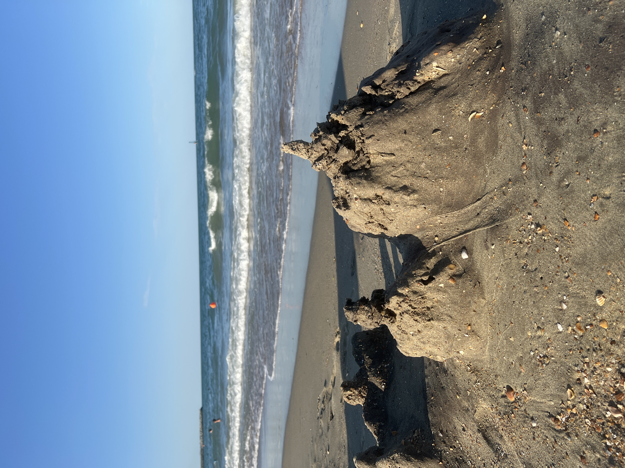 The image shows a close-up of sandcastles on a beach with the ocean in the background. The sandcastles are small and appear to be made of wet sand. They are in various stages of construction, with some being more detailed than others. The ocean is calm with gentle waves. The sky is clear and blue. The location is the beach in Constanța, Romania. There are a few people visible in the water, and an orange buoy in the distance. The sand is littered with small shells and pebbles. The lighting suggests it is daytime, likely late afternoon due to the angle of the shadows.