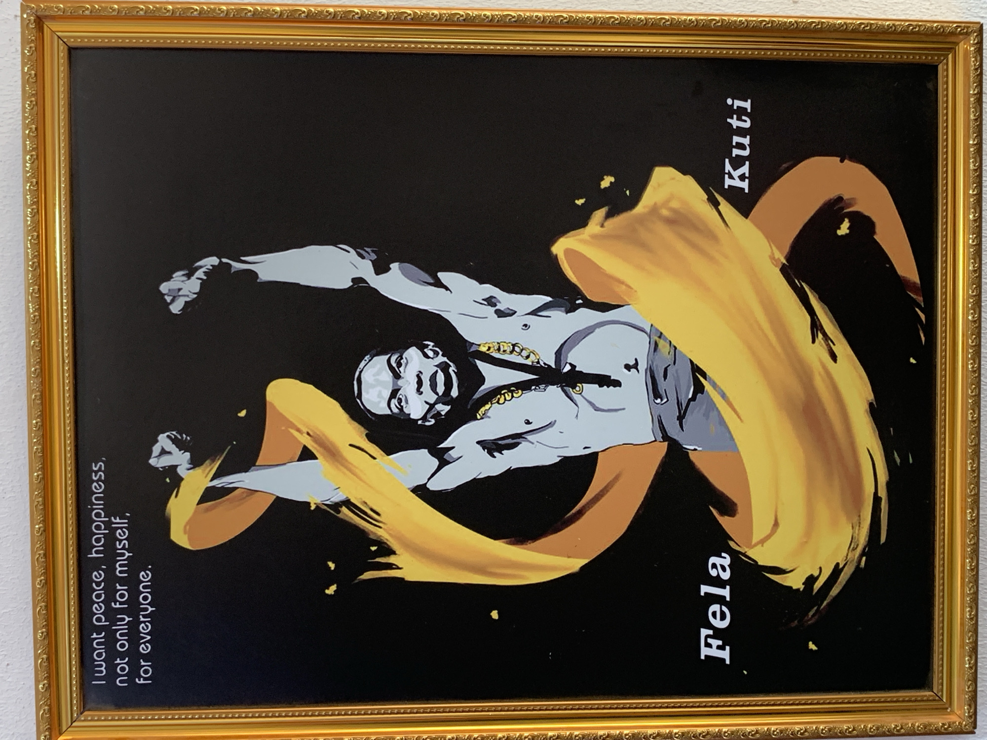 A framed portrait hangs against a light-colored wall. The frame is ornate and gold. Inside the frame is a black background on which a figure is painted. The figure appears to be a man with dark hair, wearing a gold chain around his neck. A swirling yellow and tan sash appears to wrap around his body. The name "Fela" is written vertically in white letters near the bottom right, and "Kuti" is written above it. On the left side, "I want peace, happiness, not only for myself, for everyone" is written vertically in white letters. The image is likely a reference to Fela Kuti. There is no information in the image to indicate it is located in Ikorodu, Nigeria.