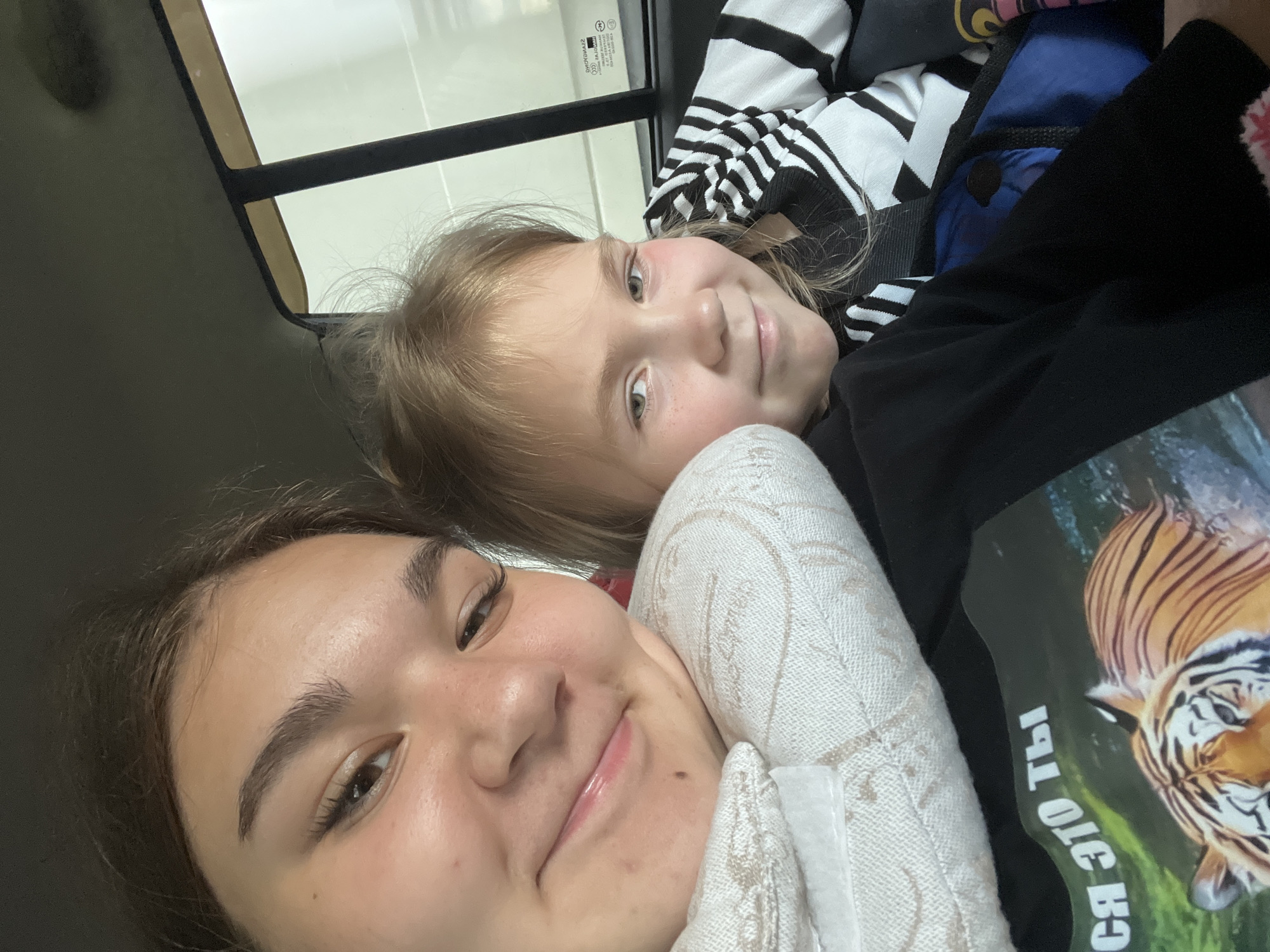 The image shows two young females in the back seat of a vehicle. The female in the foreground on the left is resting her head on a white pillow. She has dark brown hair and is looking towards the camera with a slight smile. The female to her right has blonde hair and is also looking towards the camera with a smile. She is wearing a black and white striped shirt. The background shows the interior of the car, including a window with a reflection of what appears to be a residential area or a street with buildings. The lighting suggests it is daytime. The image appears to be a selfie taken from inside the vehicle. Based on the presence of a "SSANGYONG" logo on the window, the vehicle is likely a SsangYong. However, there are no specific landmarks or visual cues to definitively place the location in Goldyrevskii, Russia. The image captures a candid moment between two individuals within a car.