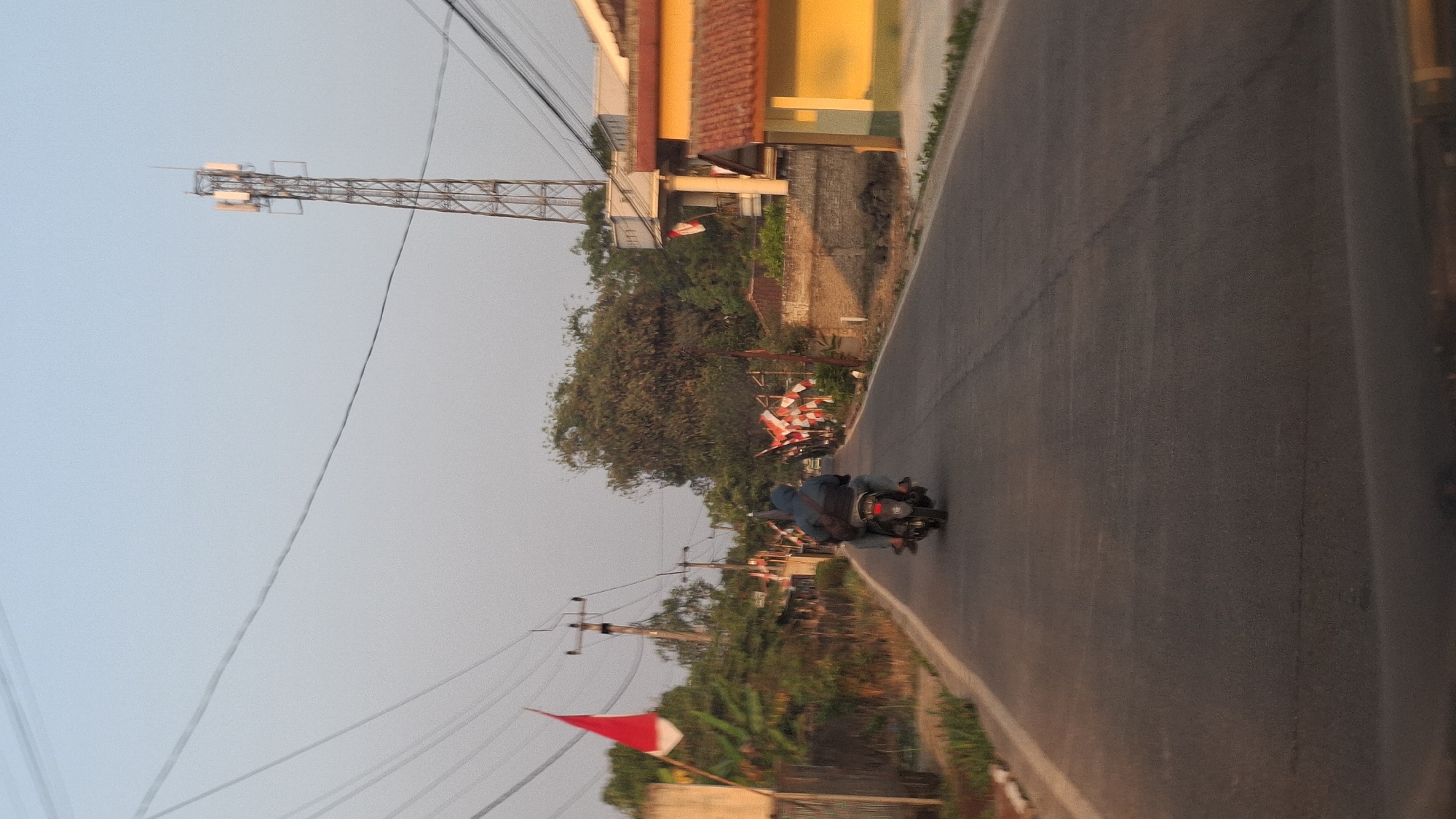 The image depicts a street scene in Cibinong Hilir, Indonesia, likely captured during daylight hours with a slightly hazy, warm sky. The primary focus is a paved road with a motorcycle carrying two passengers riding away from the viewer, moving towards the upper right of the frame. The passenger in the rear is wearing a hooded jacket.
Along the left side of the road, a red and white Indonesian flag is prominently displayed on a pole. Further down the road, on both sides, there are numerous smaller red and white Indonesian flags arranged along the roadside, suggesting a local celebration or event.
Vegetation, including trees and bushes, lines the sides of the road, interspersed with houses. A tall telecommunications tower is visible in the background on the right side of the frame, along with buildings that appear to be residential or commercial structures, one of which is painted yellow. Overhead, power lines traverse the sky, supported by utility poles. The overall atmosphere is one of a rural or suburban Indonesian community.