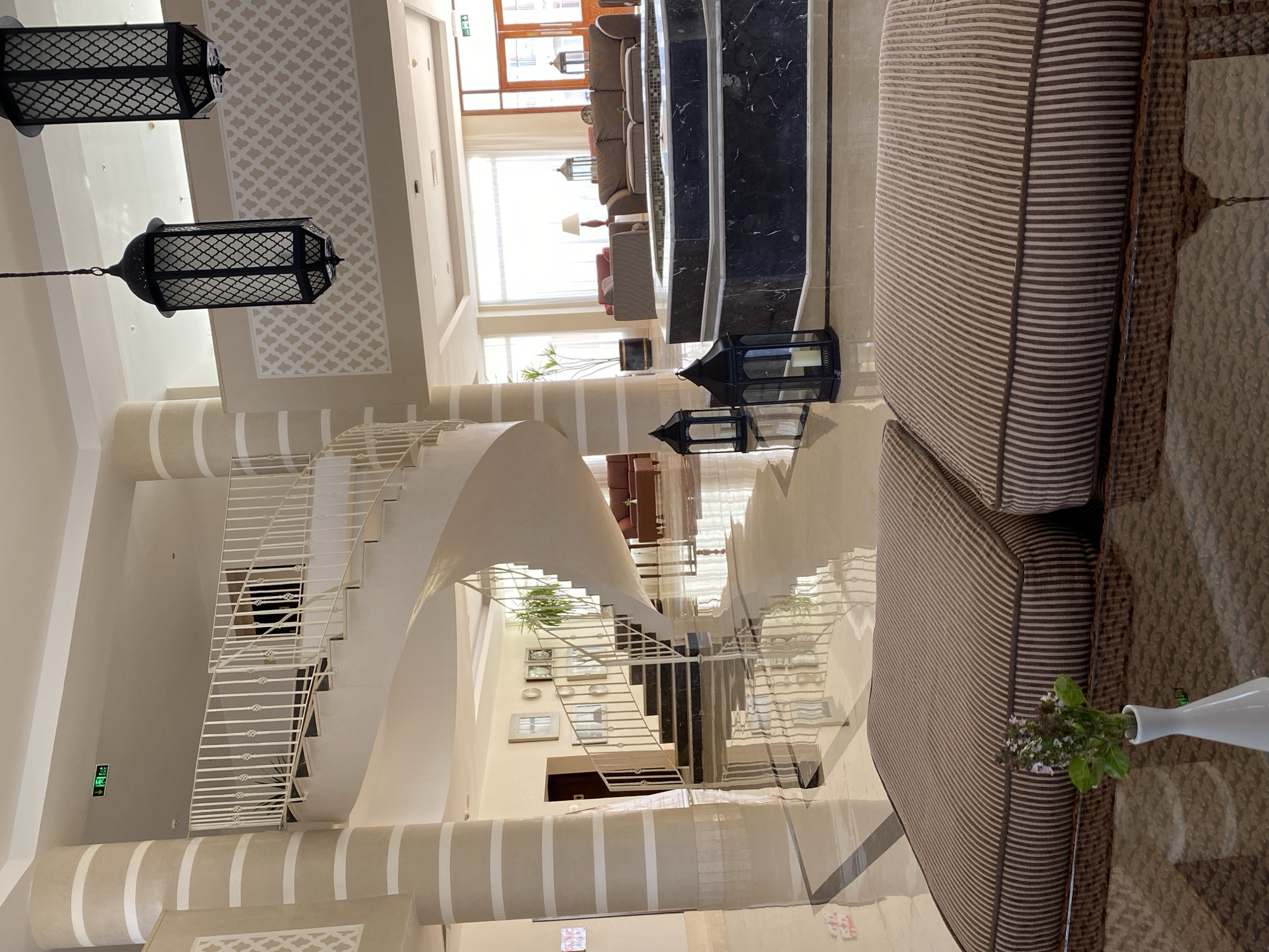 The image depicts the interior of a hotel lobby, likely in Sharm El Sheikh, Egypt, characterized by its spacious design and elegant decor. Dominating the scene is a grand, white spiral staircase with ornate railings, a central architectural feature. To the left, large, cream-colored columns with beige horizontal stripes add a distinct visual element. The floor is highly polished and reflective, mirroring the staircase and surrounding architecture.
In the foreground, two plush ottomans with brown and beige stripes are visible, suggesting a seating area. A small, white vase containing a sprig of green foliage sits on a glass-topped coffee table in front of the ottomans. Further into the lobby, a dark marble fountain with a decorative black railing is present. Around the fountain, several black, lantern-style lamps with intricate detailing are placed, adding to the ambiance.
In the background, a seating area with comfortable-looking sofas and armchairs is arranged near a large window that allows ample natural light to enter. Two decorative hanging lanterns, also in a dark, intricate design, are suspended from the ceiling. An "EXIT" sign with green lettering is visible on the ceiling in the distance, indicating a safety feature. The overall environment suggests a luxurious and well-maintained hotel lobby, designed for comfort and aesthetic appeal. There are no people visible in the image.