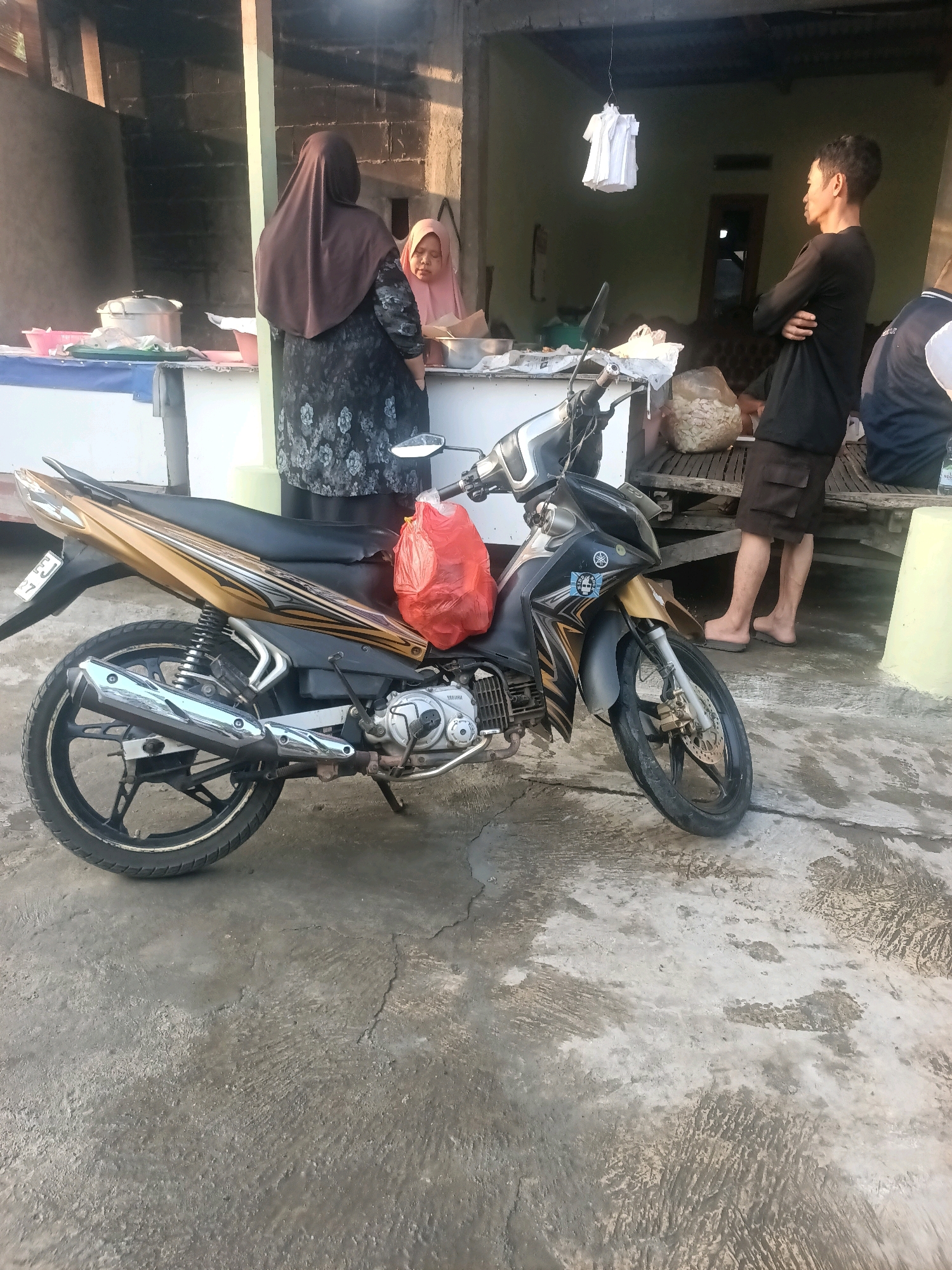 The image depicts an outdoor scene at a food stall in Wanajaya, Indonesia. A black and gold Yamaha motorcycle is parked prominently in the foreground, with a red plastic bag hanging from its handlebars. Behind the motorcycle, two women are visible at the stall. One woman, wearing a brown hijab and a patterned dress, faces away from the camera. The other woman, in a pink hijab, is partially visible. A young man in a black shirt and shorts stands to the right, with his arms crossed, facing away from the camera. The stall itself appears to be a temporary structure with a wooden counter and a thatched roof. In the background, a simple dwelling with a doorway is visible. The ground is paved with concrete.