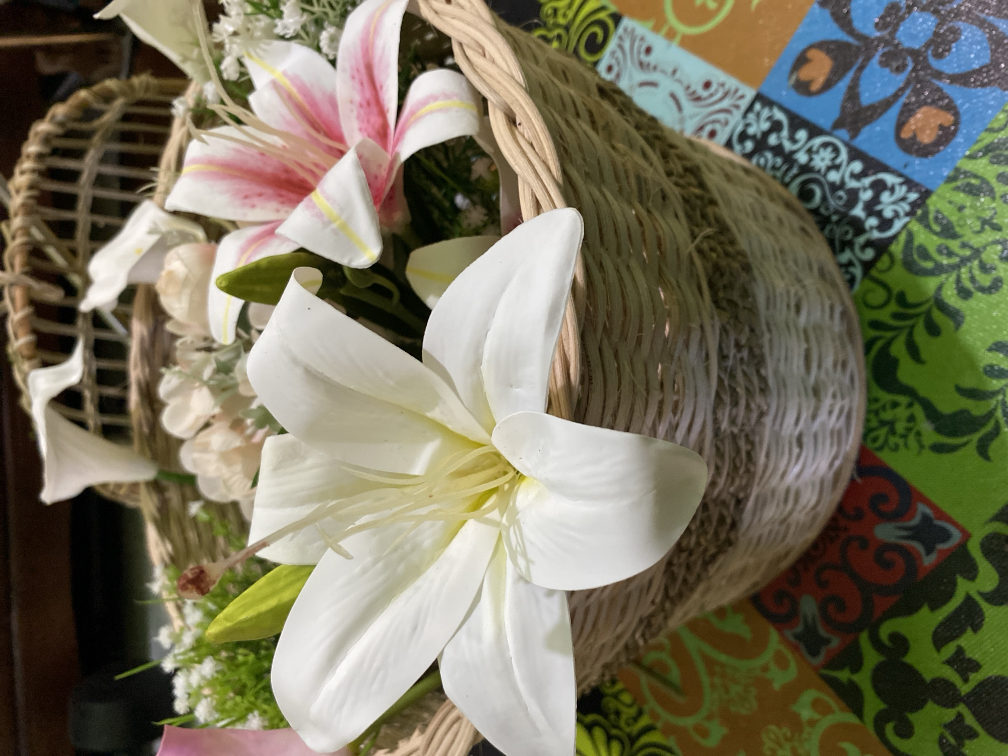 A close-up, eye-level shot shows a wicker basket filled with artificial flowers, including large white lilies and smaller pink and white lilies, along with green foliage. The basket itself is woven with natural fibers and sits on a table with a colorful, patterned surface. The background is blurred, showing what appears to be dark wood furniture. The scene is indoors, possibly in a home or a shop, and there are no people or animals visible. The lighting suggests artificial light. The image is sharp and focused on the floral arrangement.