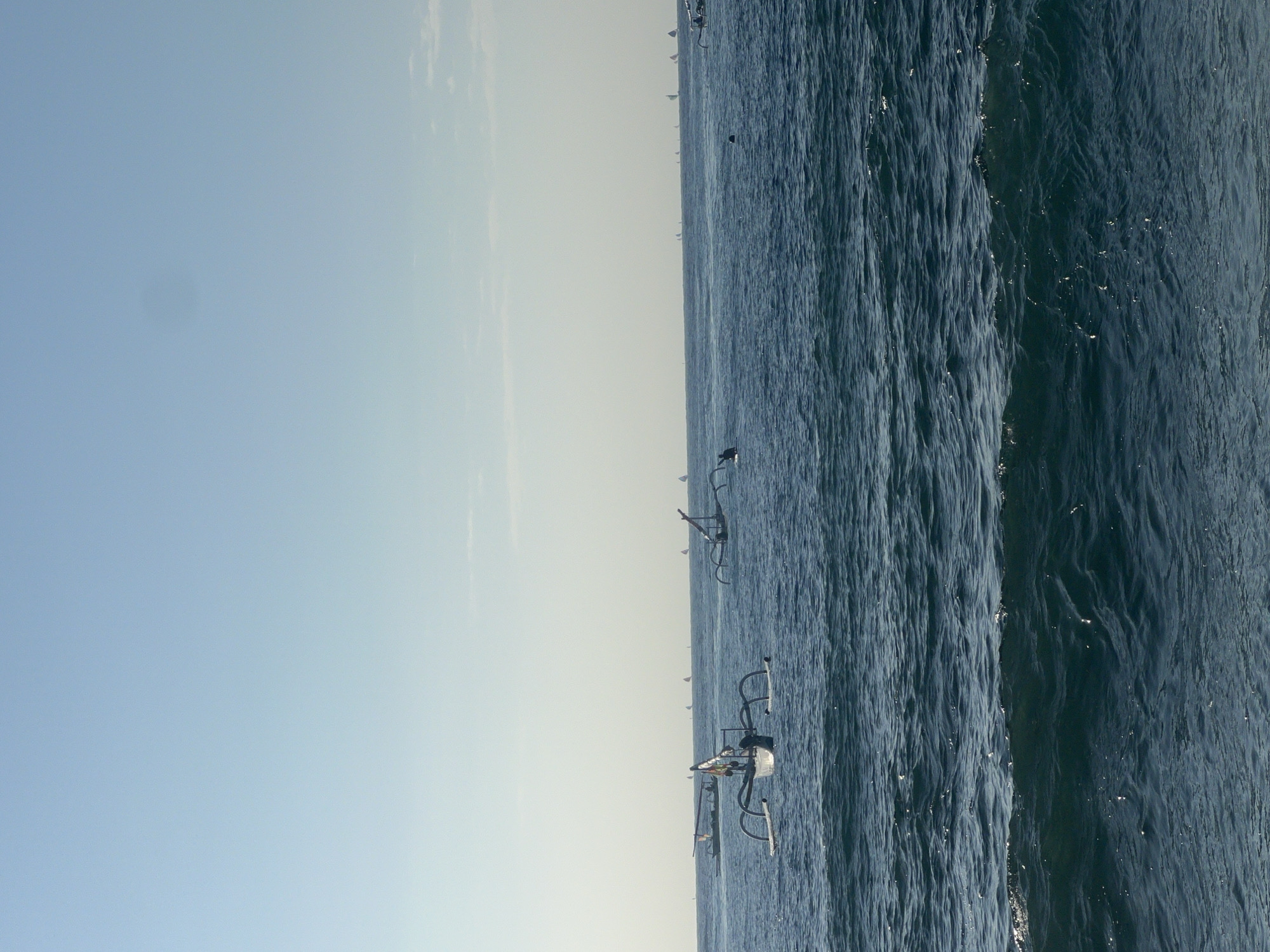 The image captures a serene seascape with a clear blue sky and calm ocean waters. Several traditional outrigger boats, known as jukung, are visible on the water. In the foreground, a single jukung with its distinctive outriggers is prominently featured. Further in the distance, more jukung boats are scattered across the horizon, along with several sailboats. The scene suggests a peaceful day at sea, likely in a coastal area like Abang, Indonesia, known for its fishing villages and traditional boat culture. The lighting indicates it is either early morning or late afternoon.