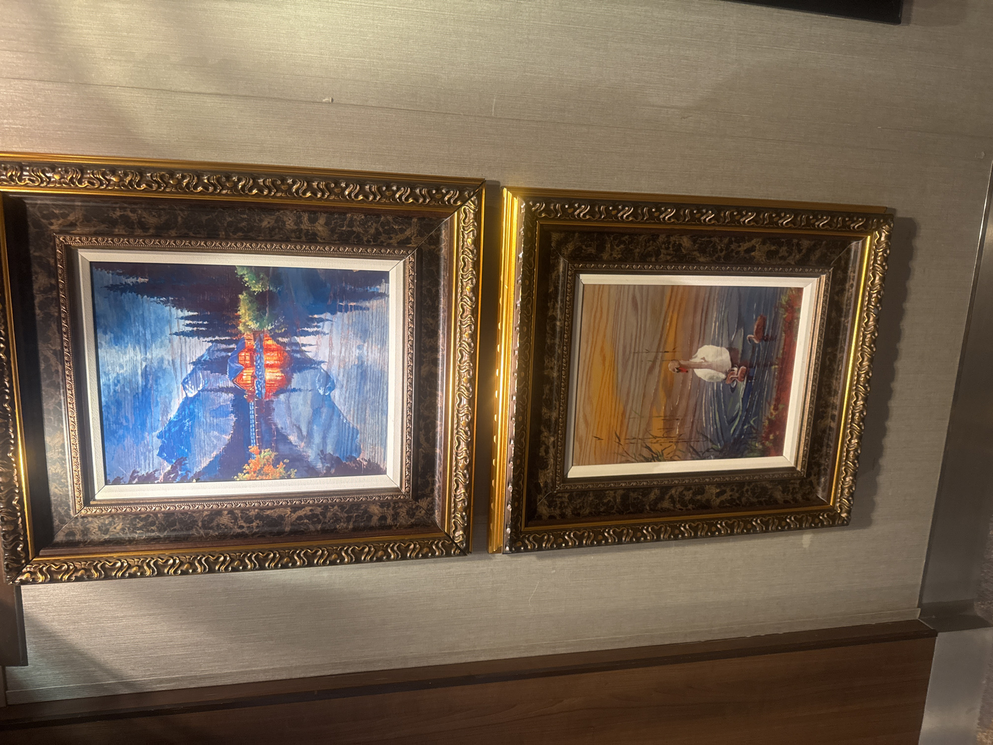 Two framed paintings hang on a textured wall. The top painting depicts a mountainous landscape with a body of water in the foreground, reflecting a cabin and trees illuminated by warm light. The bottom painting shows a swan and cygnets on water, with reeds in the background. Both frames are ornate with gold and dark brown accents. To the left of the paintings is a wooden door frame. The location in the city of Motril, Spain is not discernible from the image.