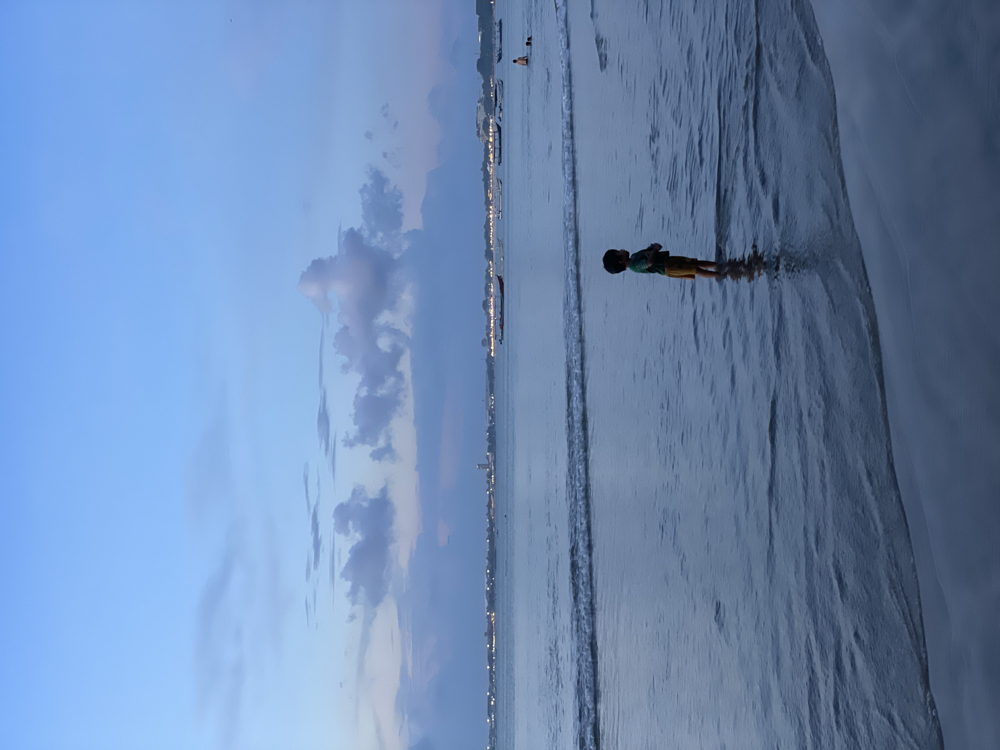 The image captures a serene coastal scene at dusk or twilight, rotated 90 degrees counter-clockwise from its intended landscape orientation. The sky transitions from a soft blue at the top to darker blues and purples closer to the horizon, dotted with scattered, light-grey clouds.
In the foreground, a young boy, wearing a green shirt and yellow shorts, stands facing right in the shallow water. Small waves gently break around his ankles, reflecting the fading light. His figure is silhouetted against the slightly lighter water.
Further out in the calm, dark blue sea, a faint line of distant lights stretches across the horizon, indicating a town or city along the coastline. Several indistinct boats are visible on the water near these lights, with other individuals wading or swimming in the distance, appearing as dark silhouettes. The right side of the image (when correctly oriented) shows the sandy, wet shoreline, marked by the retreating tide. The overall atmosphere is peaceful and calm, characteristic of an evening at the beach.
No visible text is present in the image. Given the location context, this scene likely depicts a beach in Kuta Selatan, Indonesia.