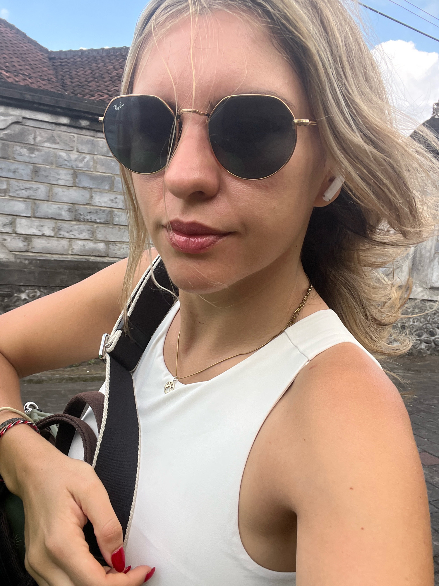 A woman with blonde hair and sunglasses poses for a selfie outdoors. She is wearing a white tank top and a gold necklace. A bag strap crosses her chest. The background shows a brick wall and a tiled roof, suggesting a building in the city of Kediri, Indonesia. The sky is visible with some clouds.