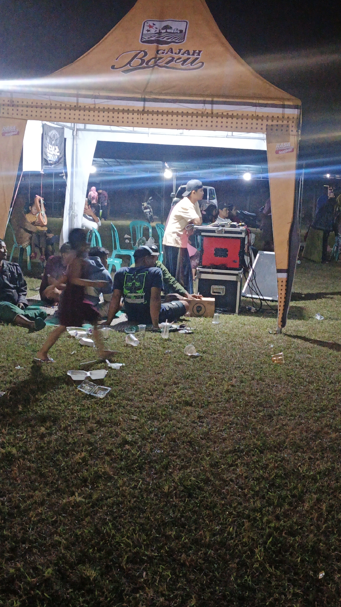 The image depicts an outdoor gathering under a large, tan tent with "GAJAH BARU" written on it. The event appears to be taking place in Ketemasdungus, Indonesia, as indicated by the tent's branding. Several individuals are present, including a child in a maroon dress running in the foreground, a person in a light-colored t-shirt and a baseball cap operating audio equipment, and others sitting on the grass. The ground is covered in grass and littered with plastic cups, food containers, and other debris. In the background, there are more tents, chairs, motorcycles, and people. The lighting suggests it is evening or night.