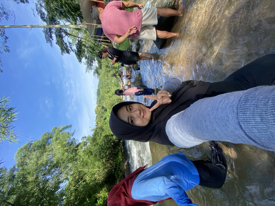 A group of people are enjoying time at a waterfall. One person, wearing a black hijab and gray sweater, is taking a selfie, holding up a peace sign. Others are wading in the shallow water behind them. The surroundings are lush with greenery and a clear blue sky is overhead. The water appears shallow and flowing. Some people are fully clothed.