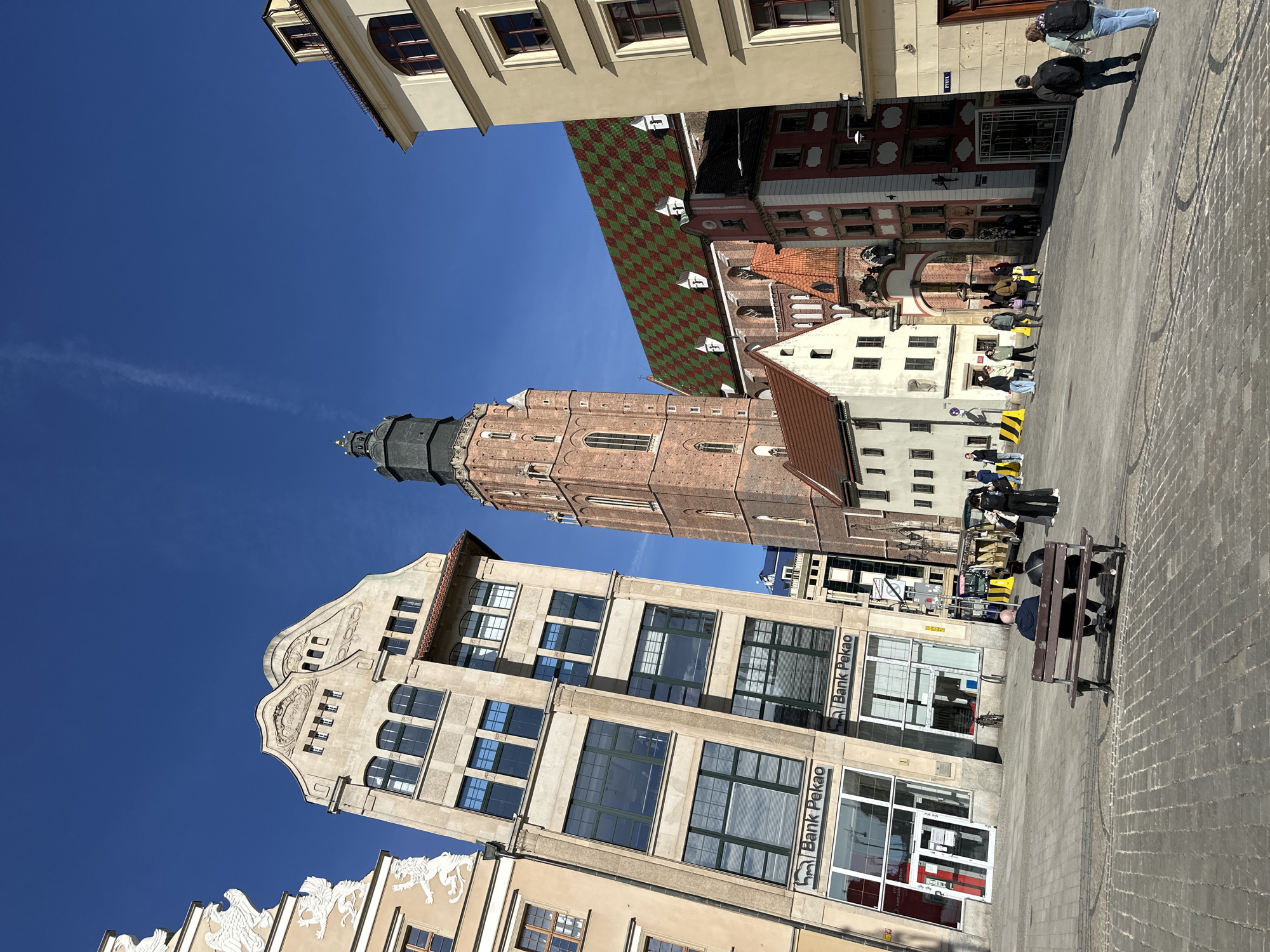 The image captures a street scene in Wrocław, Poland, under a clear, blue sky. Dominating the view is a tall, brick church tower with a dark-colored spire, rising between other buildings. One of the structures is a modern building with large windows and signage for "Bank Pekao" visible on its facade. The architecture is a mix of historical and contemporary styles. In the foreground, a section of the paved street is visible with a person sitting on a bench. Pedestrians walk along the edge of the buildings in the midground. A building with a green and red checkered pattern roof is seen on the upper middle right.