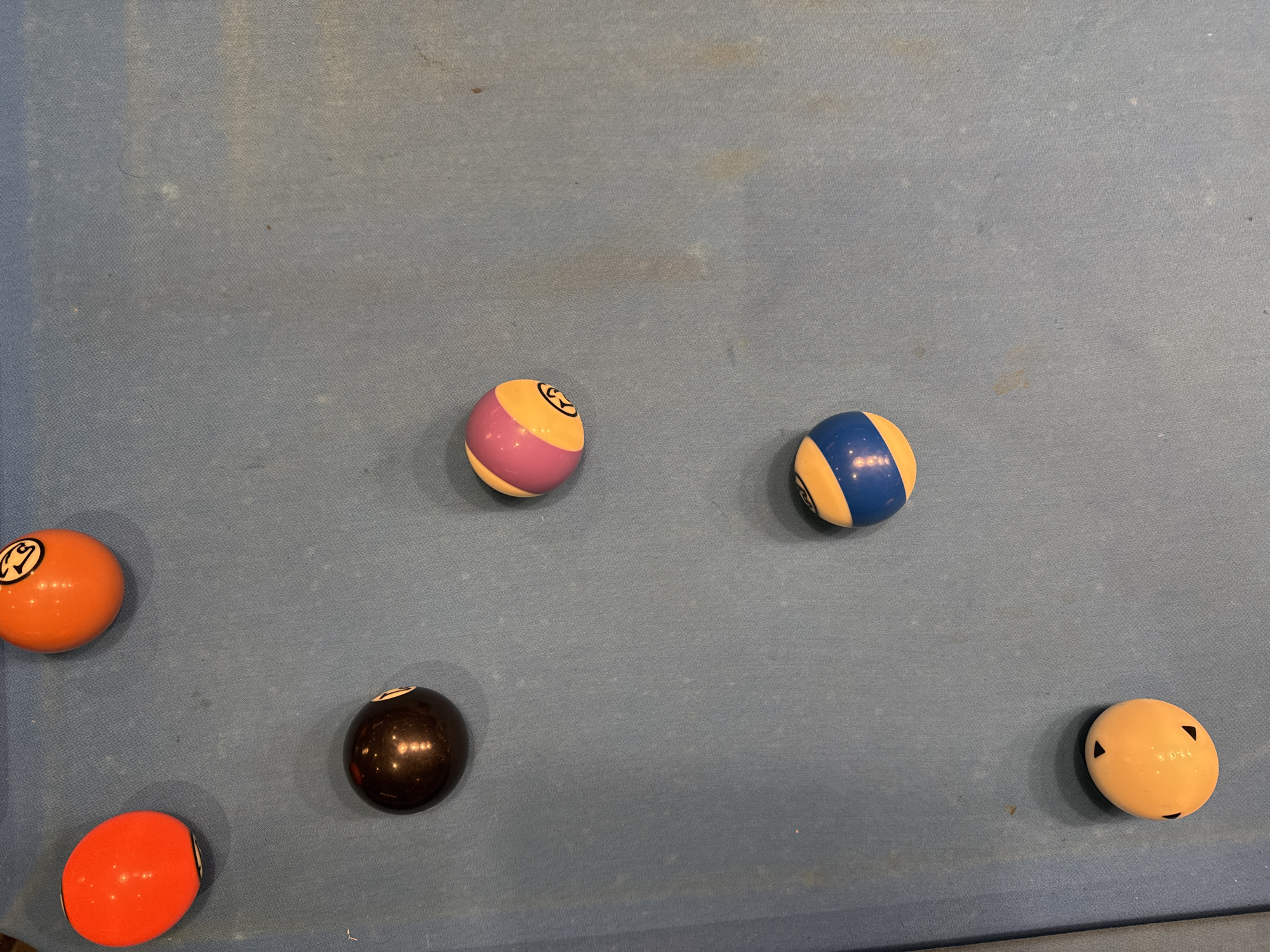 The image depicts a blue pool table with six pool balls scattered on the surface. The balls are: an orange 13-ball, a purple/yellow striped ball, a blue/yellow striped ball, a black 8-ball, an orange ball, and a red ball. The pool table shows signs of wear. The location in Bellville, South Africa is unknown.