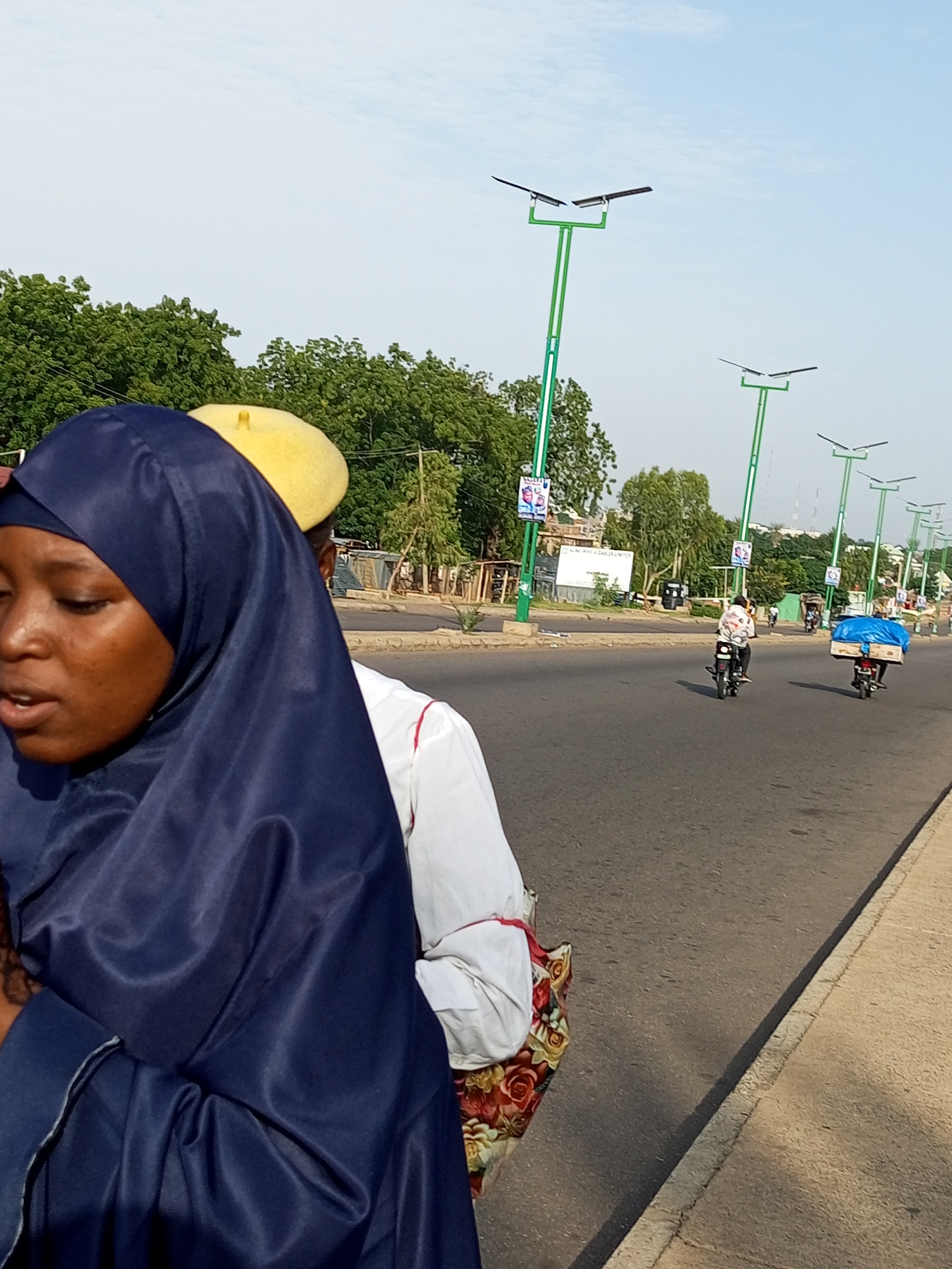 The image captures a scene on a street in Tumfure, Nigeria. In the foreground, a woman wearing a dark blue hijab is partially visible, with her face turned to the left. Behind her, a person wearing a bright yellow cap is also visible. They appear to be on a street with a paved road and a sidewalk. Several streetlights with solar panels line the road, which is busy with traffic. Two motorcycles are prominent on the road: one in the middle distance with a rider wearing a patterned shirt, and another further back with a rider carrying a large blue tarp-covered load. Trees and buildings are visible in the background, under a clear sky.