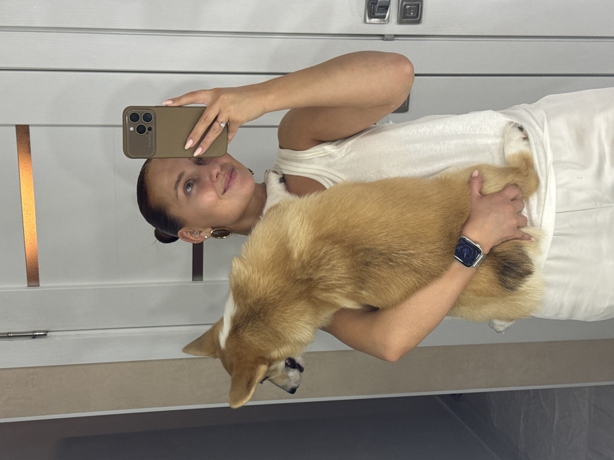 A woman is holding a corgi dog while taking a selfie with a phone. She is wearing a white top and a smartwatch. The background appears to be an interior setting with a door. The location is Chaikovsky, Russia.