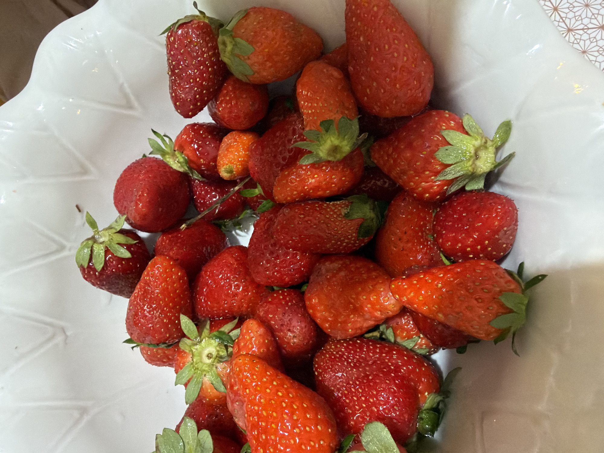 A close-up shot shows a white, possibly decorative, bowl filled with numerous fresh strawberries. The strawberries are various sizes and shades of red, some with attached green stems. The bowl sits indoors, near a surface with a geometric pattern. There is no evidence to confirm this image was taken in Budva, Montenegro.