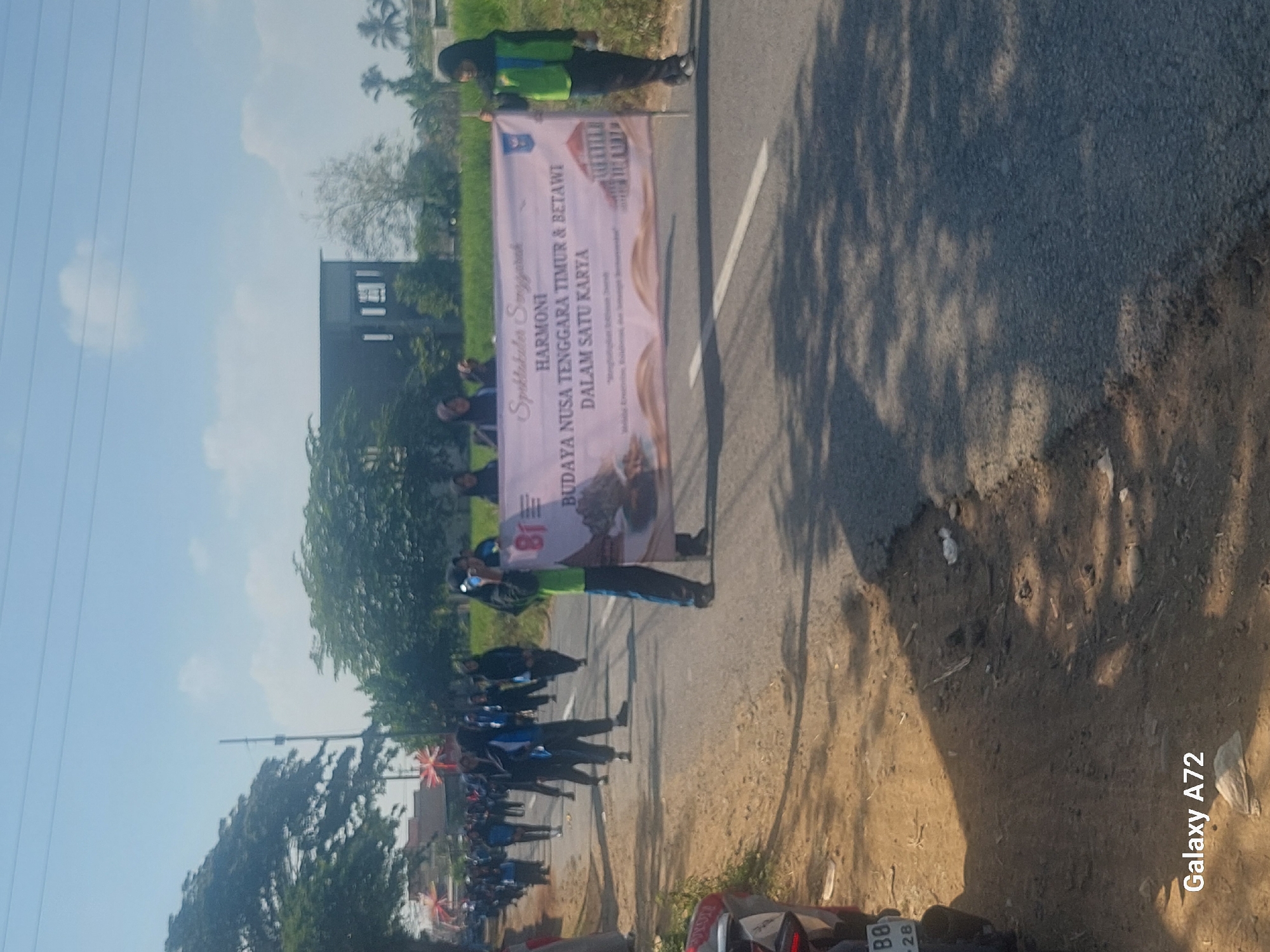 In Sengguruh, Indonesia, a group of people, mostly young students wearing hijabs and uniforms, are participating in a parade or procession on a sunny day. They are walking along a paved road, with trees and buildings visible in the background. The students are carrying a banner that reads "Spektakuler Sengguruh HARMONI BUDAYA NUSA TENGGARA TIMUR & BETAWI DALAM SATU KARYA". Some of the students are wearing green vests, and one person in the foreground is holding a camera and taking pictures. A vehicle with the license plate "BB 28" is parked on the side of the road. The ground is covered with dirt and shadows from the trees.