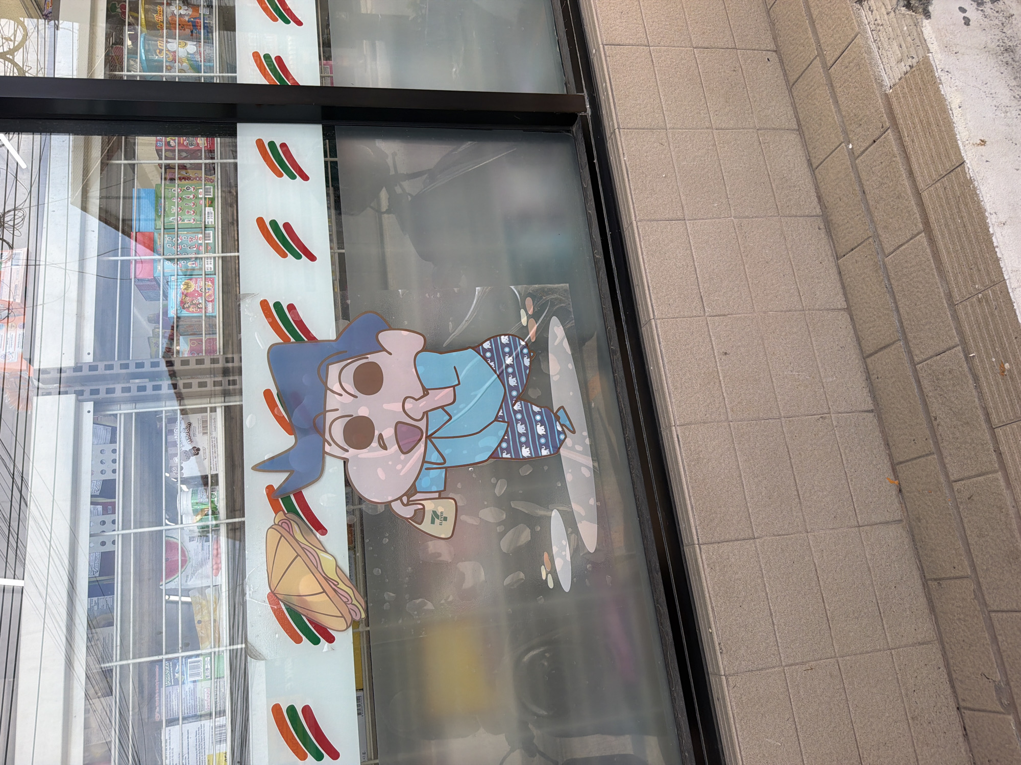 The image shows a display window of a 7-Eleven store in Baan Mae Nam, Thailand. The window is decorated with a cartoon sticker of a character resembling Shin-chan, a popular anime character. The sticker depicts the character wearing a blue shirt and patterned pants, holding a 7-Eleven shopping bag and standing near a sandwich. The window also has the 7-Eleven logo with its characteristic three stripes. Behind the sticker, shelves stocked with various products are visible, including what appear to be snacks and drinks. The exterior of the store features a tiled facade.