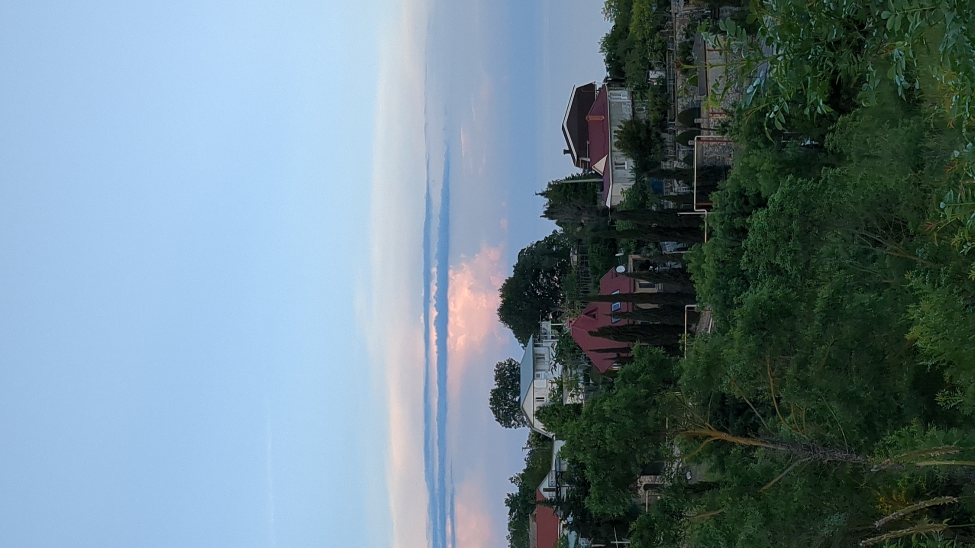 The image shows a view of Kiparisnoe, Ukraine, featuring several residential buildings with red roofs interspersed with trees. The sky is partially cloudy with vertical cloud formations exhibiting shades of blue and pink. Lush green foliage is prevalent in the foreground and around the houses. The composition is vertically oriented.