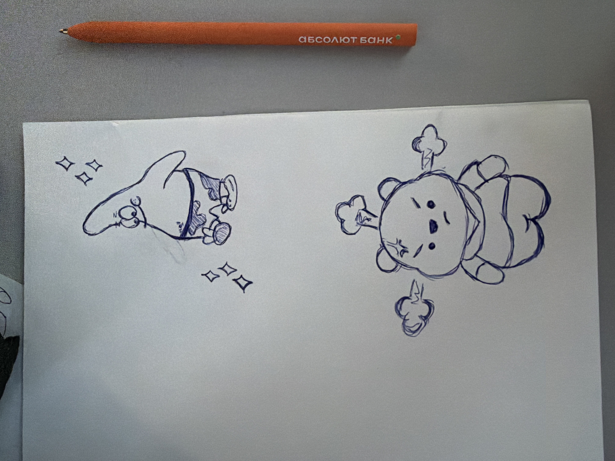 A close-up shot shows a sheet of white paper with blue ink drawings of two cartoon characters and a bright orange pen with text in Russian on the right side. The upper drawing depicts a character resembling Patrick Star from SpongeBob SquarePants, with his characteristic body shape, eyes, and shorts. Above him are four small, stylized diamond shapes, suggesting sparkle or magic. The lower drawing shows a character resembling Winnie the Pooh, with a grumpy expression and puffs of smoke or steam emanating from his sides, indicating anger. The pen is positioned vertically, its textured surface visible, and the Russian text on it reads "АБСОЛЮТ БАНК". The background is a neutral gray surface. The image appears to have been taken in an indoor environment, possibly a desk or table. There is no discernible indication of the specific location within Rostov-on-Don, Russia.