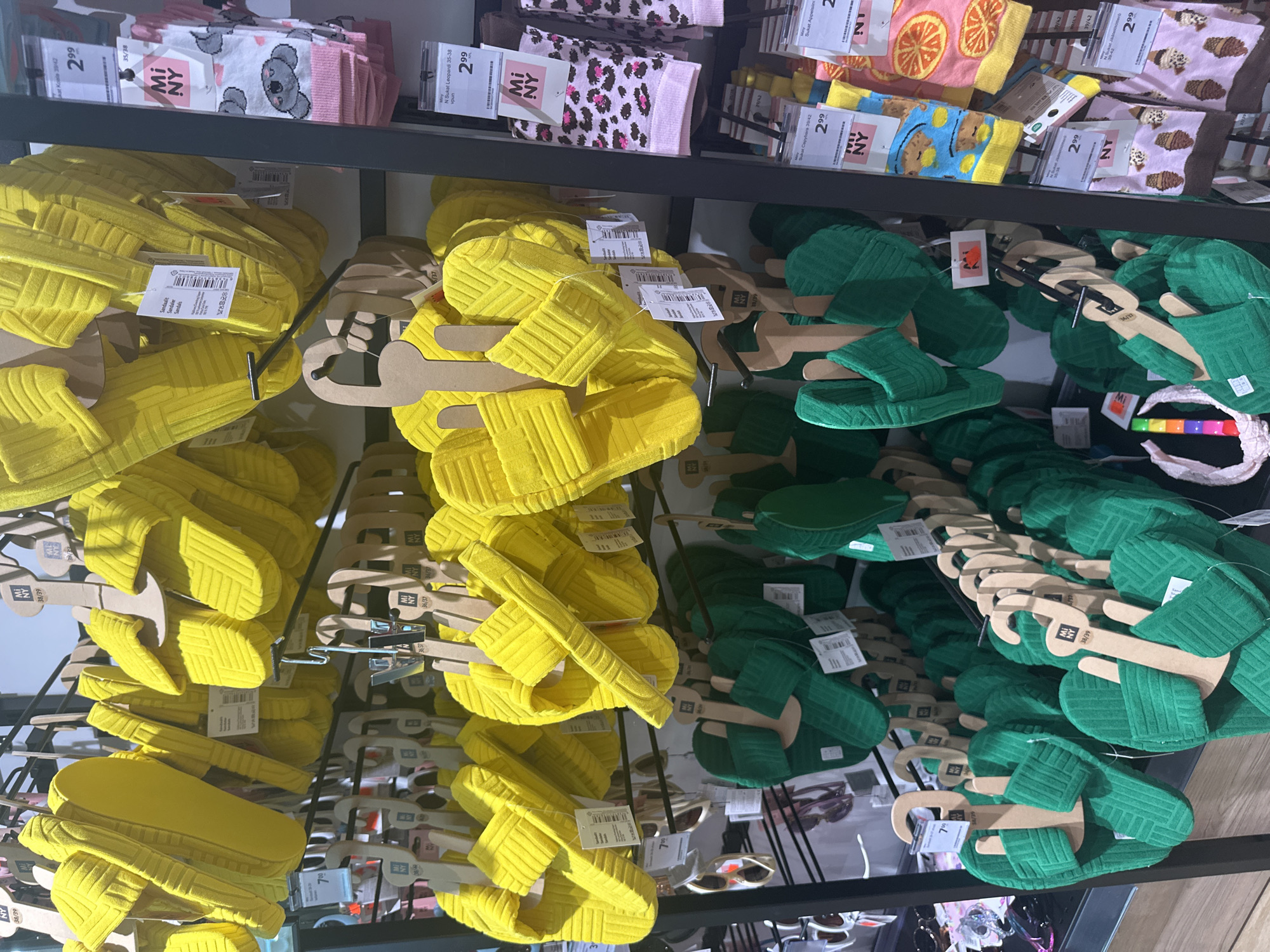 This media file shows a retail display of footwear, specifically slippers. The slippers are organized on shelves, with two primary colors visible: bright yellow and dark green. Both colors of slippers are presented in a similar style, appearing to be slip-on sandals with a textured design. They are displayed on cardboard hangers, with some hangers indicating sizes such as "38/39" and "39/42".
Above the slippers, on the top shelf, is a display of socks. These socks feature various colorful and playful patterns, including koalas, leopard print, oranges, and ice cream cones. Some of these socks also have price tags displaying "2.99" and branding like "Mi NY".
The overall setting appears to be a retail store, likely a department store or clothing boutique, with multiple shelves dedicated to displaying these items. The lighting suggests an indoor environment, typical of a commercial space. There are no people visible in the image. The focus is solely on the merchandise, showcasing the variety and presentation of the slippers and socks. The visible text primarily consists of product information such as brand names, item types, sizes, and prices.