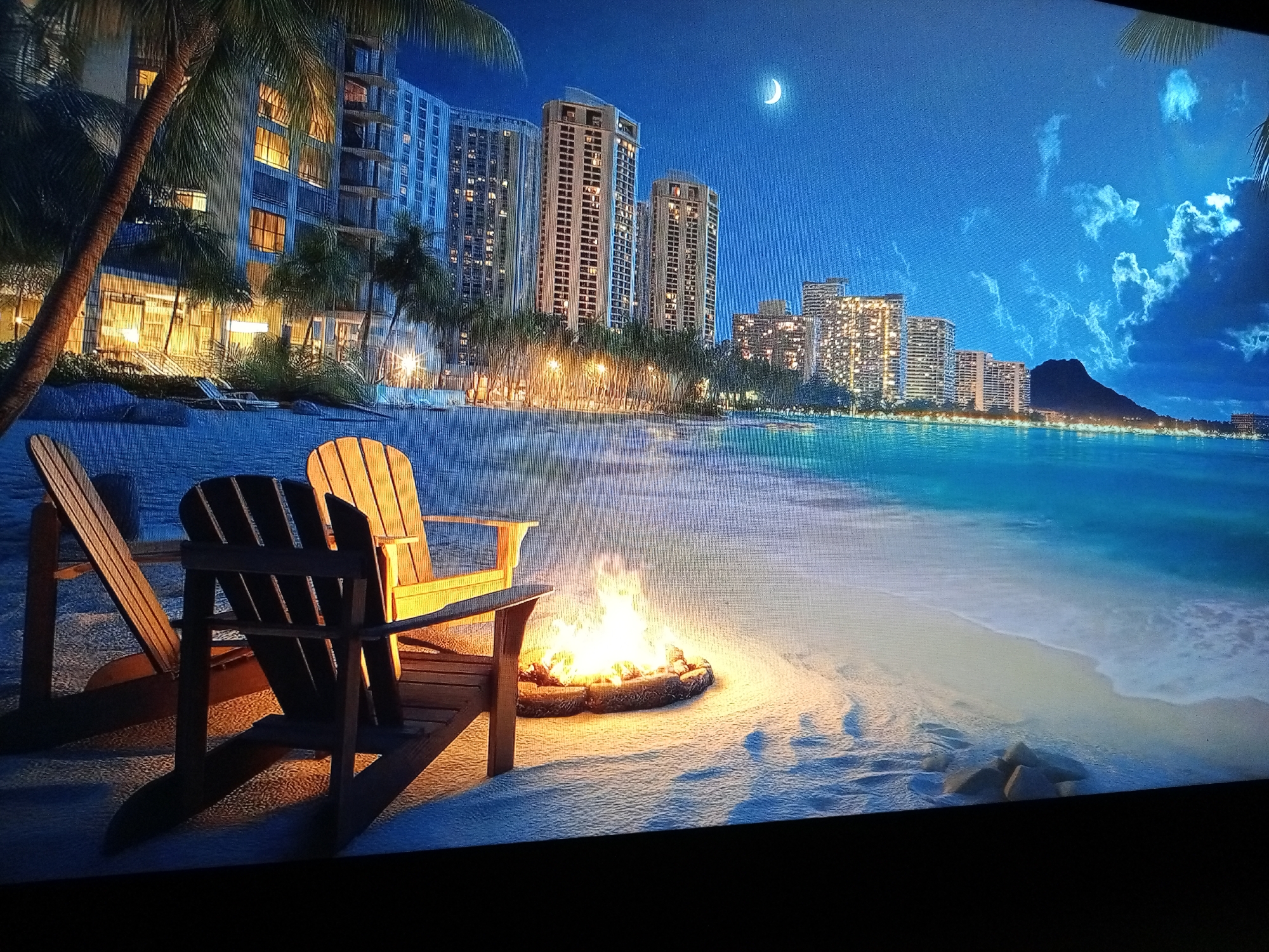 The image depicts a serene beachfront scene at night. Two wooden Adirondack chairs sit on a sandy beach near a small bonfire contained within a stone ring. In the background, the ocean meets a coastline lined with tall, illuminated buildings suggesting a resort or city skyline. A crescent moon is visible in the night sky. Diamond Head, a volcanic tuff cone, is also visible in the distance. Palm trees are present. The image does not contain any people. The location is not Tinaquillo, Venezuela; it is likely Waikiki Beach, Honolulu, Hawaii.