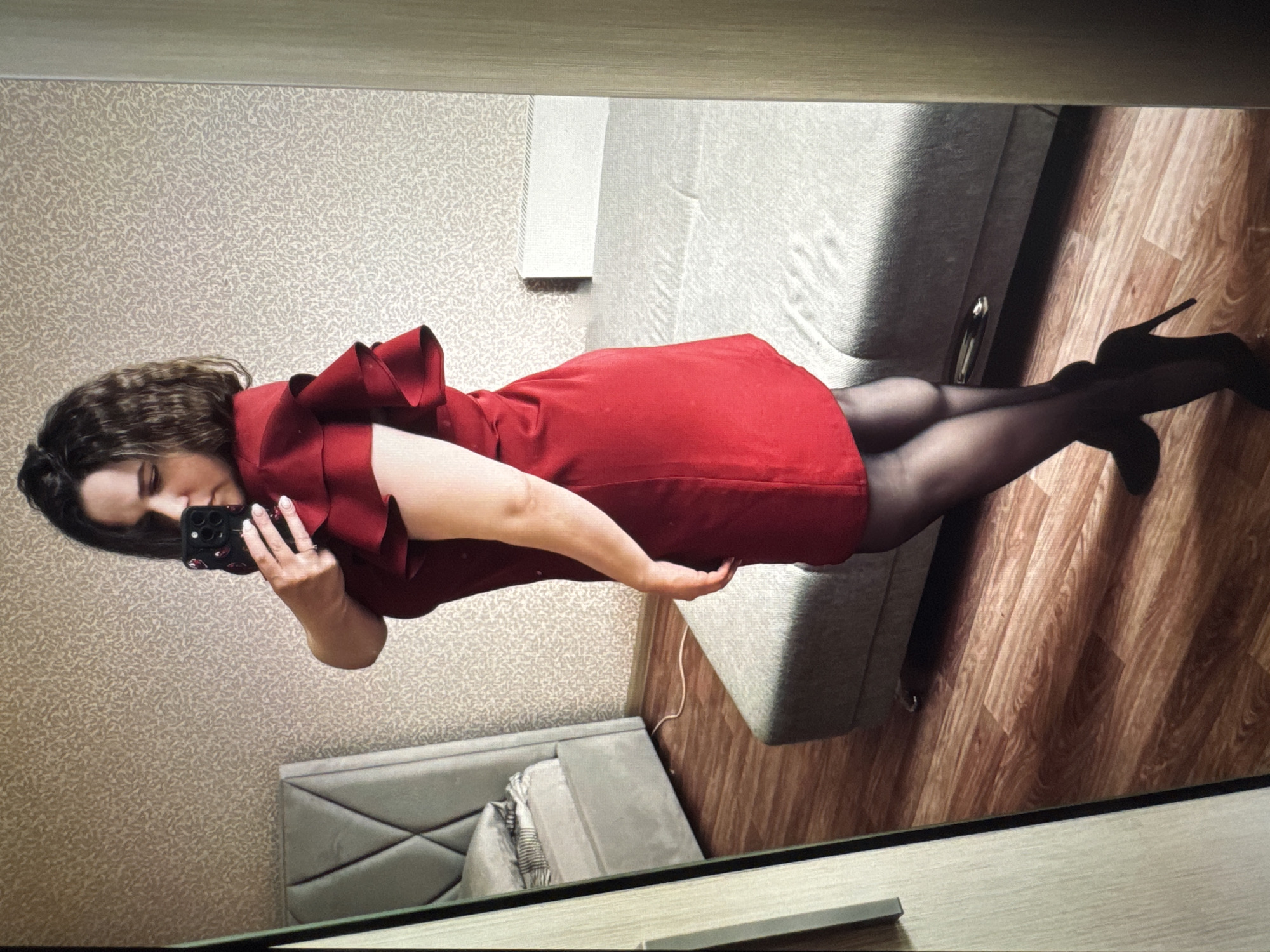 A woman is seen in a full-length mirror selfie. She is wearing a red dress with a large ruffle detail, black tights, and black high heels. She is holding a smartphone in her left hand, presumably to take the photo. The background includes wallpaper with a textured pattern, a gray upholstered headboard of a bed, a gray couch or seating area, and wood-look flooring. Based on geolocation, the location is in Новинки, Russia.