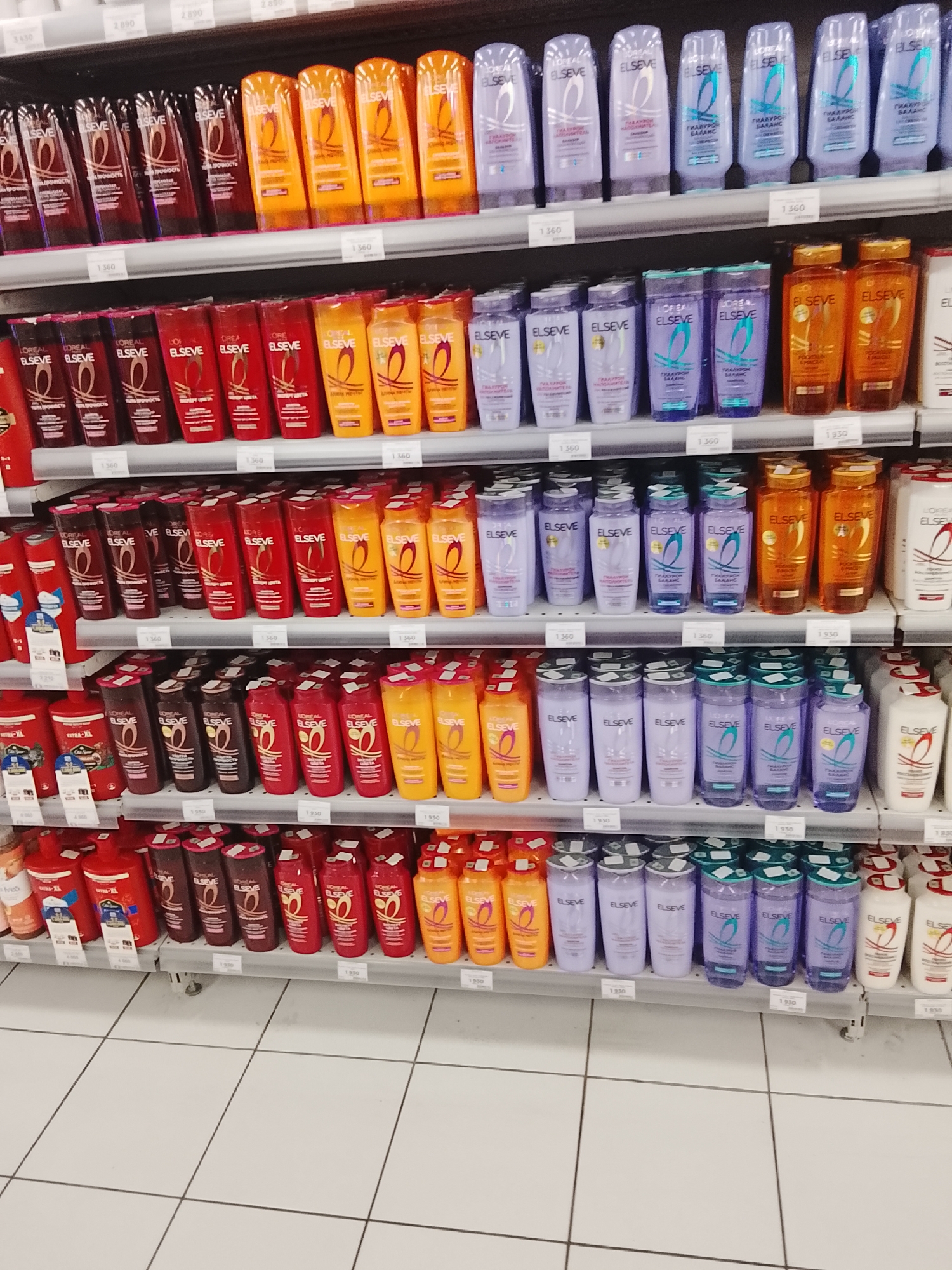 This is a retail scene, likely a supermarket or drugstore aisle, showcasing rows of personal care products. The shelves are neatly organized, displaying numerous bottles of shampoo and conditioner. The products are primarily from the L'Oréal Elseve brand, with various colors and labels indicating different product lines. Many bottles are clear, revealing colorful liquids inside, such as purple, red, and orange. Some bottles have distinct branding with the L'Oréal and Elseve logos.
Visible text includes product names, brand names, and price tags. Price tags like "1360", "1930", and "2890" are scattered throughout the shelves, suggesting these are retail prices. There are also labels with terms like "ГИАЛУРОН БАЛАНС" and "СИЛА ПРОЧНОСТЬ" in Cyrillic script, indicating the specific benefits or ingredients of the products. The lighting suggests an indoor environment, possibly under fluorescent or store lighting. The overall impression is one of abundance and variety of consumer goods. There are no people visible in the image, and the scene is static, implying a typical retail display.