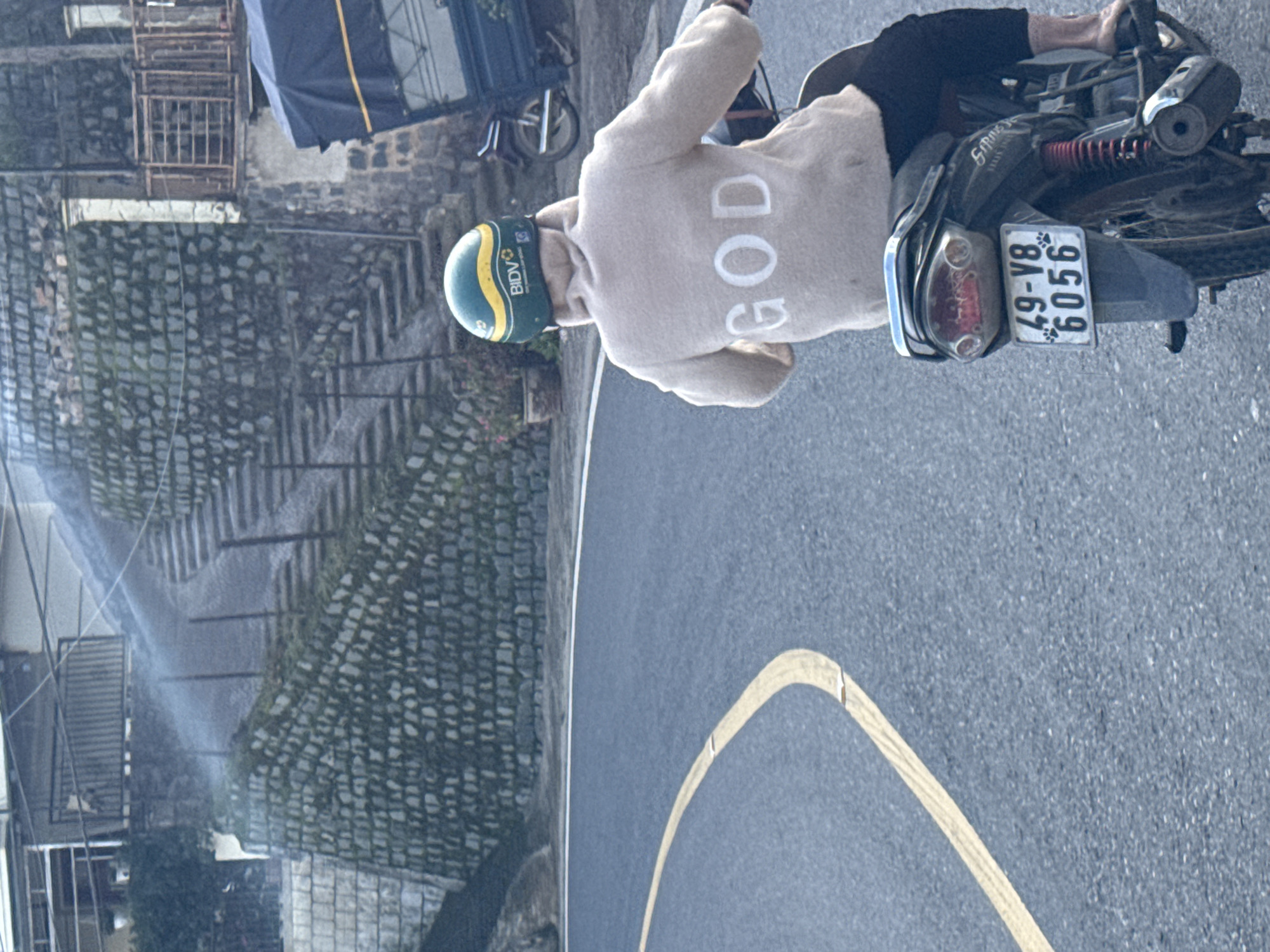 A person wearing a beige hooded sweatshirt with the word "GOD" in large white letters on the back is riding a motorcycle. The person is also wearing a green, yellow, and white helmet with the word "BIDVO" visible on it. The motorcycle has a license plate that reads "49-V8 6056". The scene appears to be a street in Phát Chi, Vietnam, with a stone wall and stairs on the left side of the road. The road has a yellow painted line. The overall lighting suggests it is daytime.
