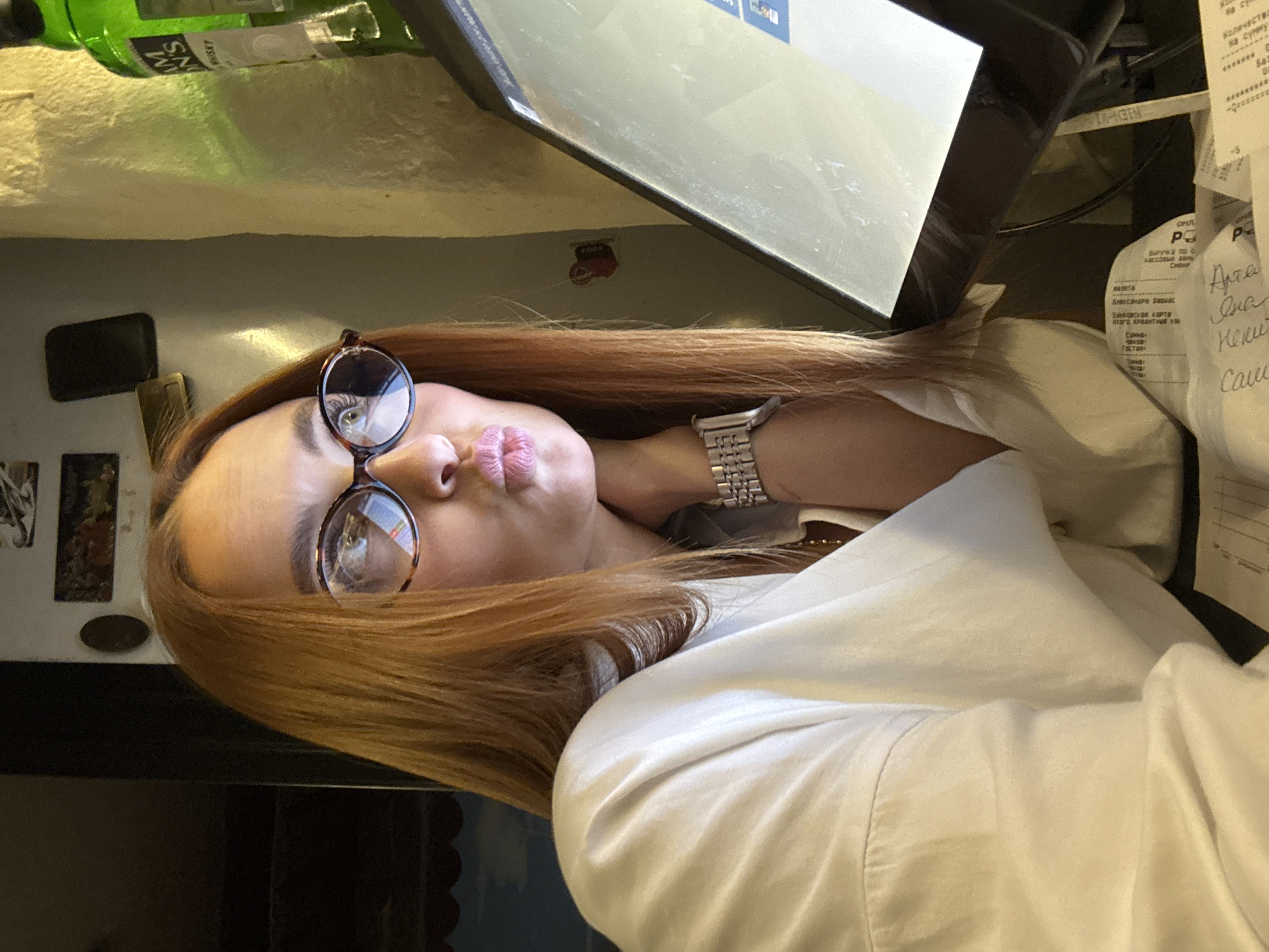 The image features a woman with long, red hair and round glasses, making a kissing face. She is wearing a white shirt and a silver watch. Behind her, there is a partially visible green bottle, a computer screen, and a receipt. The receipt contains Russian text indicating it's a transaction record, likely from a cash register, mentioning "Alexandria Davina" and "Chekhov Gastaik." Based on the OCR text on the receipt, the location is likely related to a business or establishment affiliated with Chekhov in the city of Chaikovsky, Russia.
