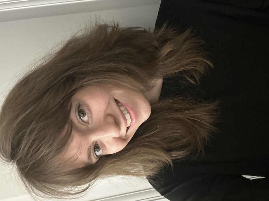 The scene depicts a young adult female, positioned indoors, likely within a residential room in Moscow, Russia. The individual has light-brown, shoulder-length hair with bangs, light-colored eyes, and a fair complexion. She is observed from the chest up, wearing a plain long-sleeved black top. The subject is smiling broadly, revealing her teeth, and maintaining direct eye contact with the viewer. Her head is slightly tilted to her left. The background consists of a plain, light-colored wall with a vertical architectural molding visible on the left side of the frame, indicating an interior setting. Lighting is even, suggesting an indoor ambient source.