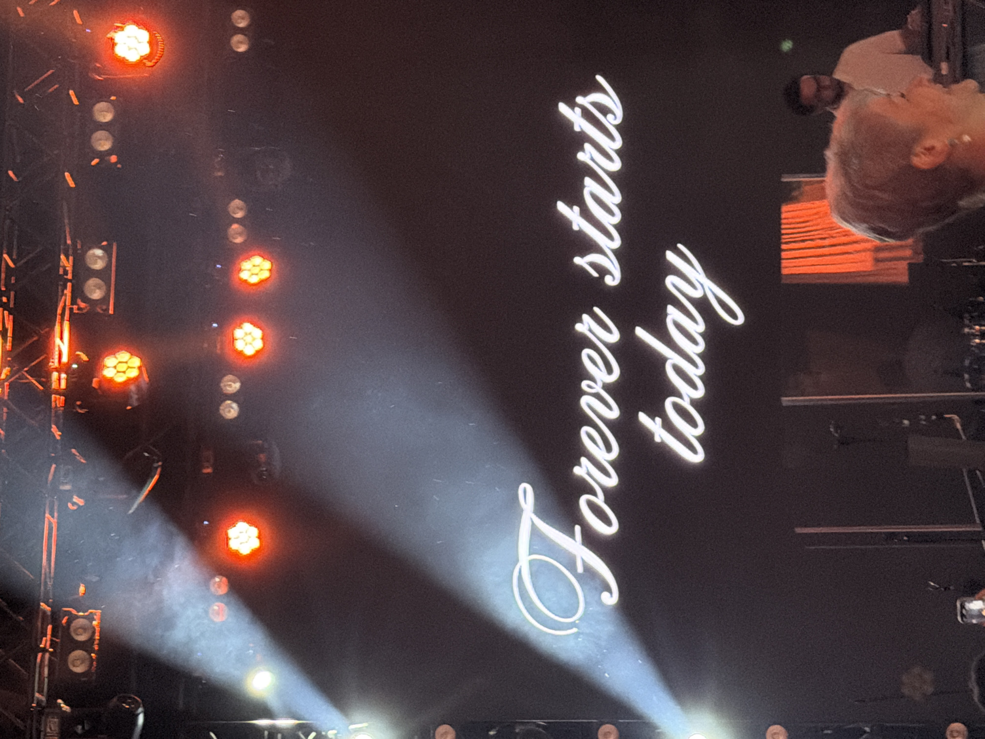 The image shows a large screen with the text "Forever starts today" written in white cursive font. The screen is illuminated by stage lights, with beams of white and orange light visible. There are several rows of stage lights above the screen, some of which are red and hexagonal. In the lower part of the image, a blurred woman with short blond hair is visible in the foreground, looking to her right and smiling. Behind her, a person wearing a white shirt is seated at a keyboard, likely a musician. The setting appears to be an indoor venue, possibly a concert hall or event space. The context of Yerevan, Armenia is not visually evident in the provided image.