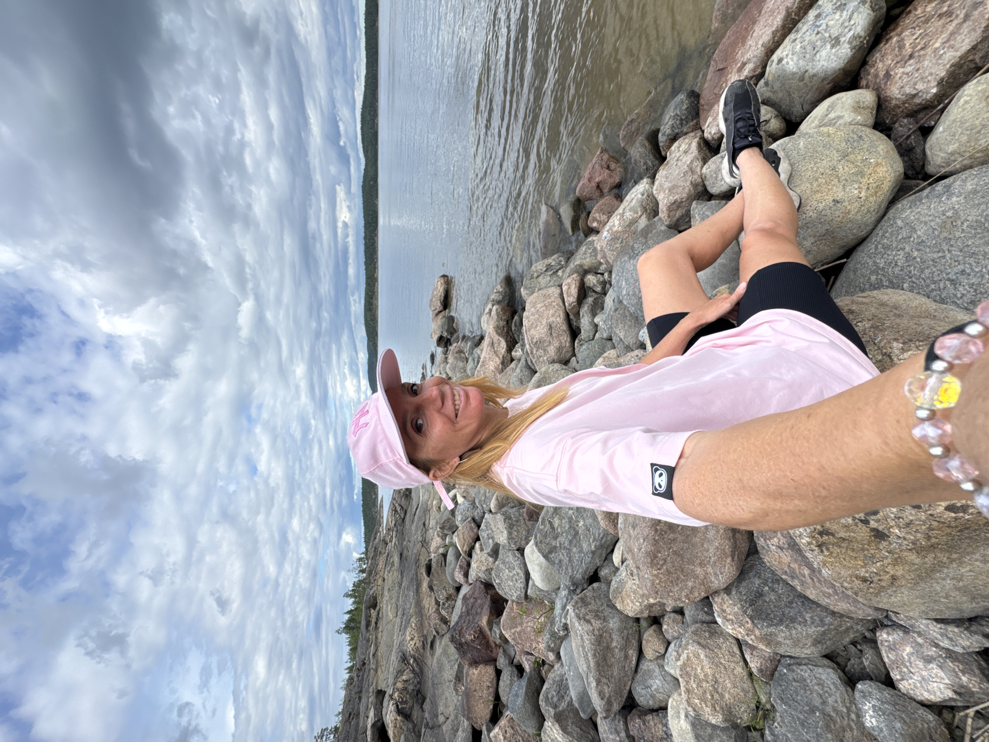 A woman with blonde hair, wearing a pink t-shirt, black shorts, and a pink baseball cap, sits on a rocky shore. She is looking at the camera and smiling. The water is calm and there are trees in the background. The sky is cloudy.