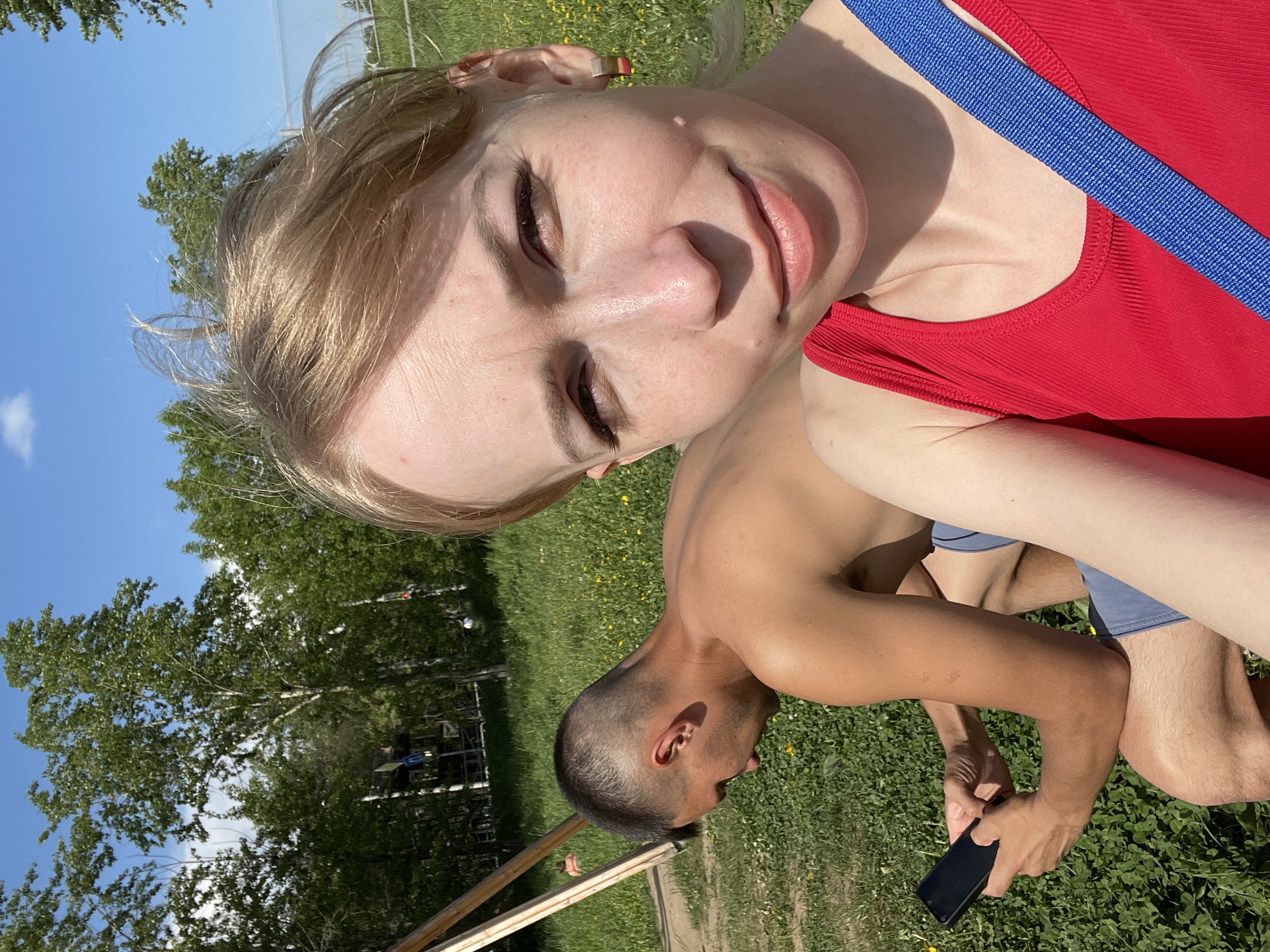 A woman in a red tank top with a blue strap is taking a selfie. A man next to her is looking down at a black smartphone in his hands. They are outside in a grassy area. Trees and a blue sky are visible in the background. The picture was taken in the city of Perm, Russia.