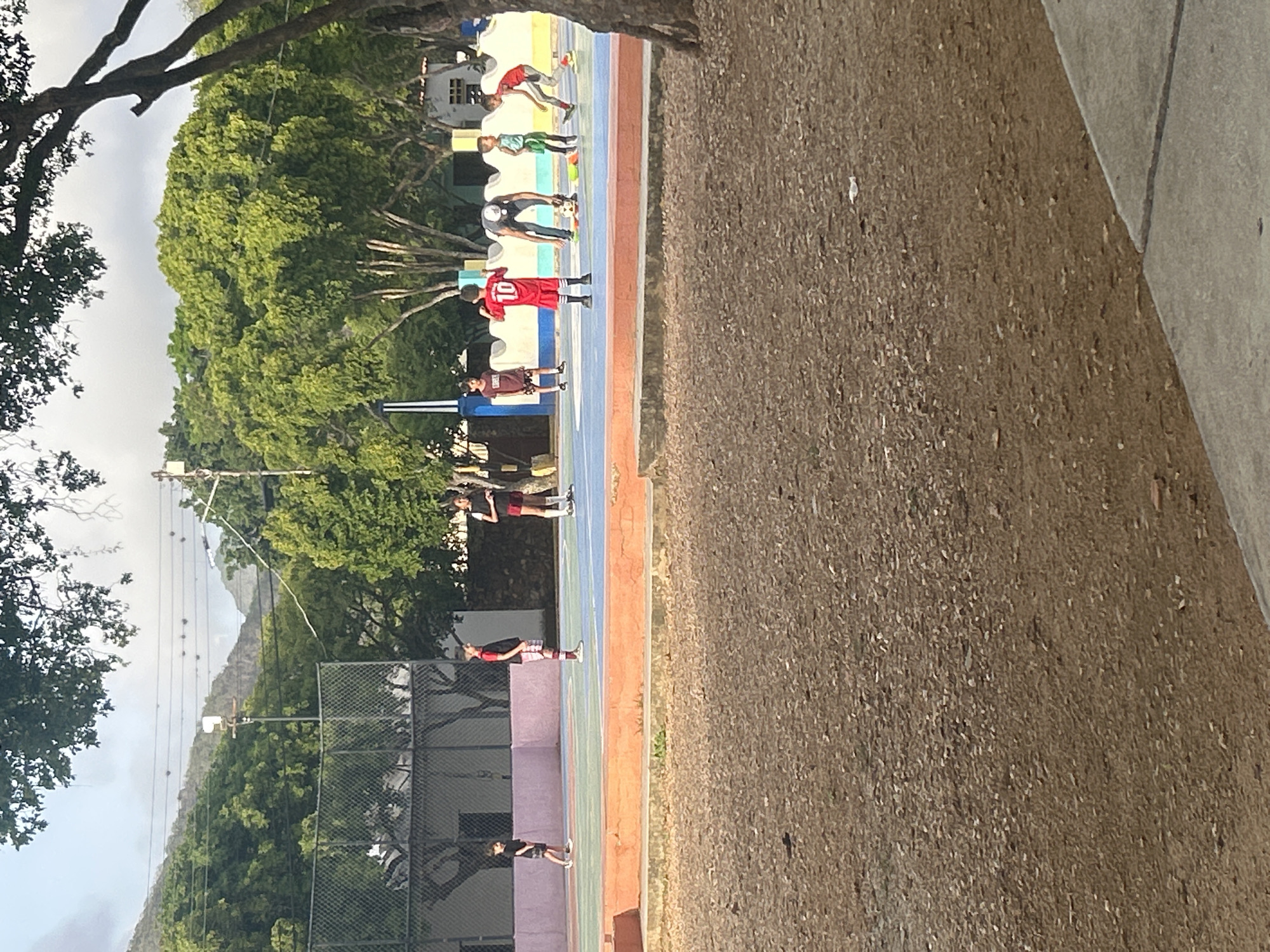 The image captures an outdoor scene in Santa Ana, Venezuela, likely at a sports complex or park. In the foreground, a patch of brown, gravelly ground is visible. Beyond this, a colorful sports court with blue, orange, and pink sections is occupied by several individuals. On the court, a group of young people, appearing to be children and adolescents, are engaged in activities, possibly a sports practice or game. One boy, wearing a red shirt with the number "10" on it, stands prominently. Another child in a red shirt and shorts is also visible, as are individuals in darker attire. A person, potentially an adult, is bent over near a soccer ball. The background features lush green trees and some buildings, with a mountainous landscape suggested in the distance under an overcast sky. A chain-link fence encloses part of the sports area.