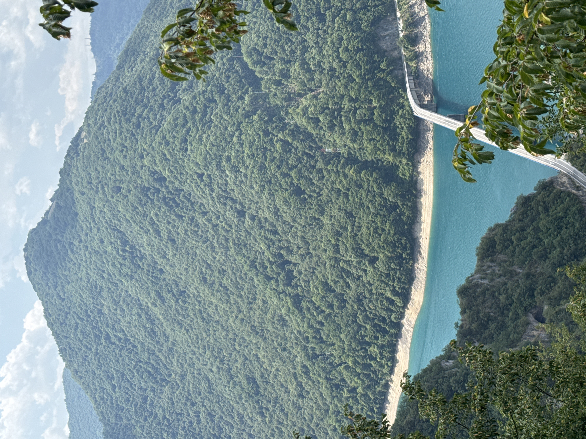 This image depicts a picturesque landscape scene near Unač, Montenegro, dominated by a vast, tree-covered mountain rising majestically. A strikingly blue body of water, likely a lake or river, carves through the base of the mountain, its shores marked by a sandy embankment. Spanning the water is a long, modern bridge, its white structure contrasting sharply with the surrounding natural elements. The bridge appears to carry a road, with vehicles subtly visible in the distance on its far end. The foreground is adorned with lush green foliage, framing the view of the mountain and water. The sky above is a soft blue, dotted with scattered white clouds, suggesting a clear, pleasant day. The overall impression is one of serene natural beauty and a harmonious blend of infrastructure with the environment.
