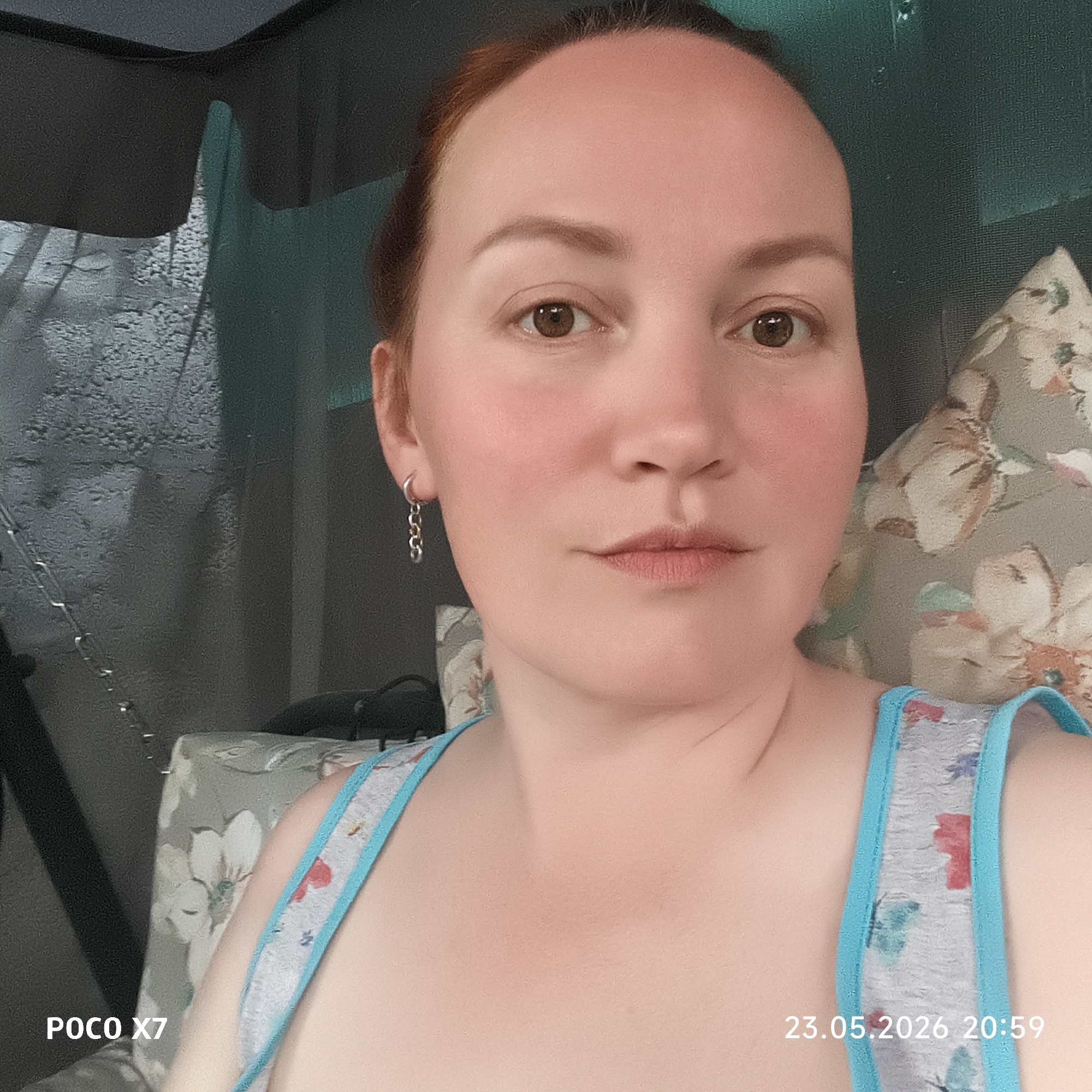 A woman with reddish hair pulled back is centered in the frame, looking directly at the camera with a neutral expression. She has brown eyes, light skin, and appears to be slightly flushed. She is wearing a light-colored tank top with a blue trim and a floral pattern. A chain earring is visible in her left ear.
She is seated on a swing or chair with floral-patterned cushions. Behind her is a semi-transparent material, possibly mosquito netting, obscuring the background. The netting appears to have moisture or raindrops on it.
In the lower left corner of the image, "POCO X7" is visible. In the lower right corner, the date and time "23.05.2026 20:59" are displayed.
The location is specified as the city of Chaikovsky, Russia.