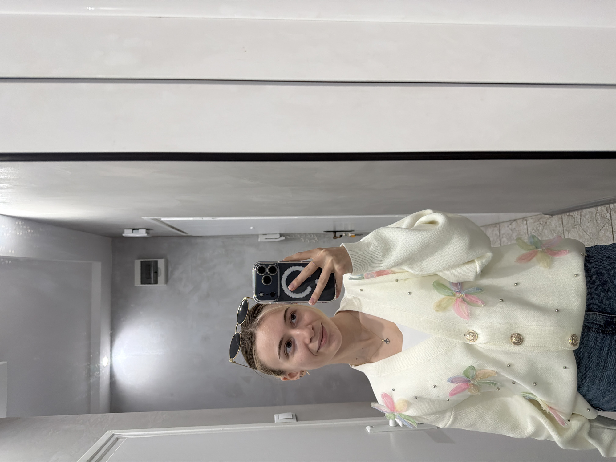 A fair-skinned young woman is taking a selfie in a bathroom or dressing room. She is holding a smartphone up at eye level. The woman has her hair pulled back and sunglasses are perched on her head. She is wearing a white cardigan decorated with pastel-colored fabric flowers and small pearl beads. A gold necklace is visible at her neck. She is wearing blue jeans.
The background shows part of a room with white and grey walls. There is a white door frame visible in the lower left corner. A light fixture and a small electrical panel are visible on the wall behind her. The location is presumed to be inside a private or commercial building, and there is no clear evidence to confirm its location in Plovdiv, Bulgaria.