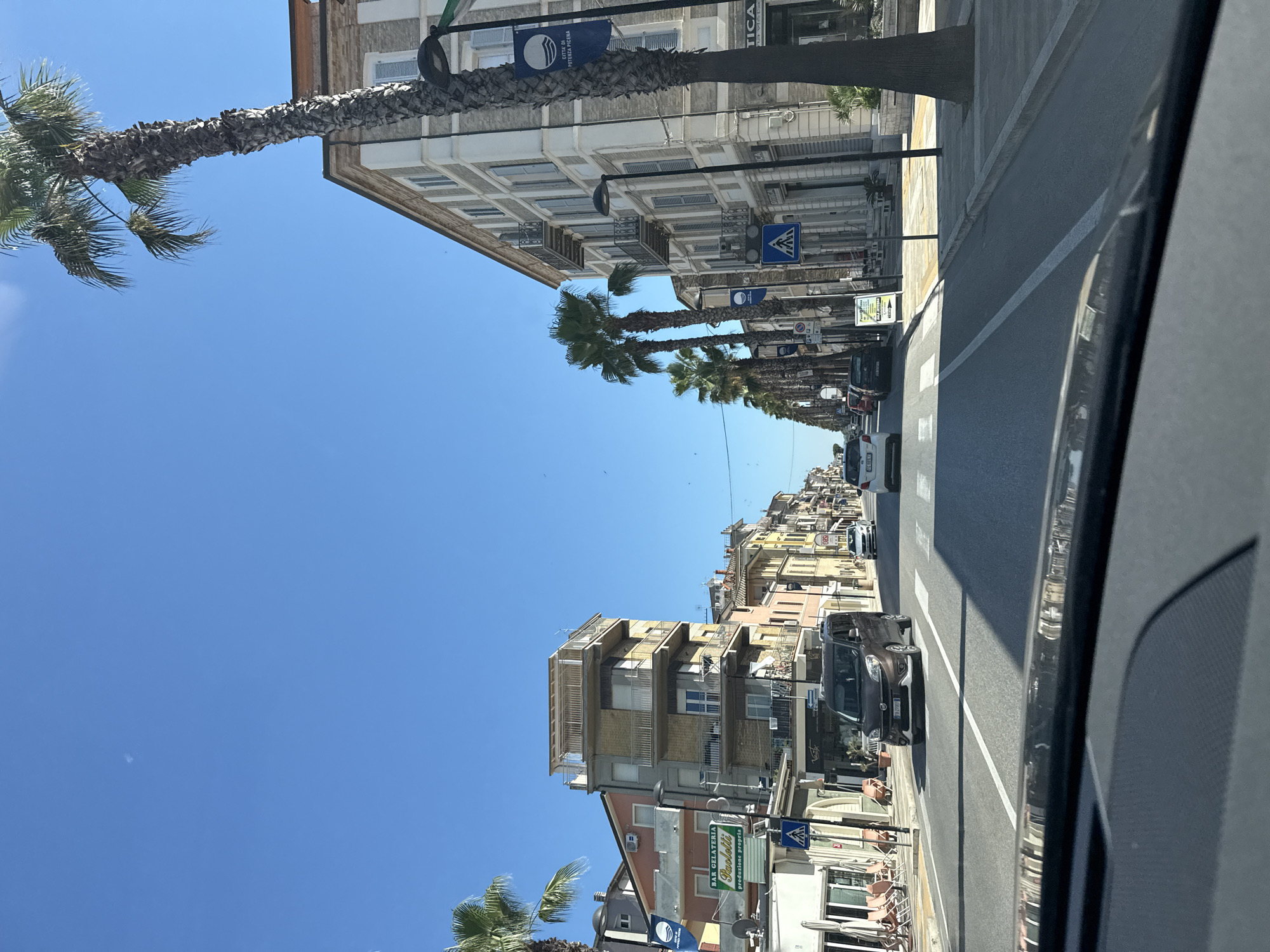 The image captures a sun-drenched street scene in Potenza Picena, Italy, viewed from inside a vehicle. The wide, paved street runs through a commercial area lined with multi-story buildings. On the right, a prominent building with light-colored stone and white-shuttered windows stands tall, adorned with a blue banner displaying a white wave logo and the text "CITTA' DI POTENZA PICENA." Several tall palm trees with rough, textured trunks line the sidewalk in front of this building. Across the street, on the left, a row of buildings in various colors, including yellow and pink, features balconies and businesses on the ground floor. One business has a green sign with white text reading "BAR GELATERIA Paolotti." Pedestrians are visible on the sidewalks, and several cars are driving on the road, including a brown Fiat Doblo minivan in the foreground. The sky above is a clear, bright blue, with a faint, small white object visible in the distance.