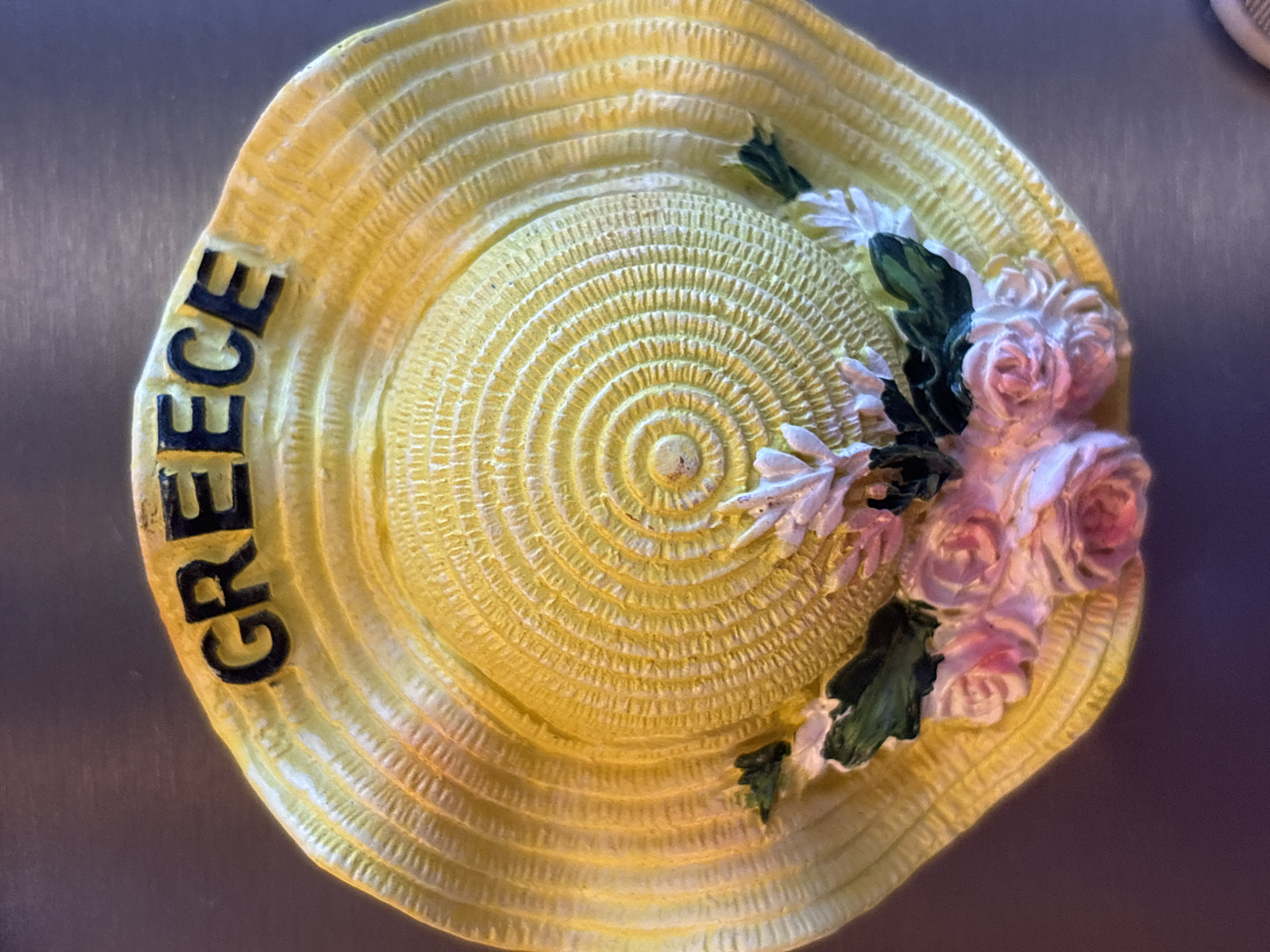 A yellow, textured hat souvenir with the word "GREECE" in black lettering is displayed against a dark, metallic background. The hat has a raised, concentric circular pattern on its crown and a brim decorated with molded pink roses and green leaves. The word "GREECE" is positioned along the top curve of the hat's brim. The lighting casts shadows and highlights, emphasizing the texture of the hat and the details of the floral embellishments. The item appears to be a decorative piece, possibly a magnet or a collectible, and its presentation suggests it is on a flat surface, such as a refrigerator or a shelf. No people are present in the image. The location is not specified in the image or the provided context.