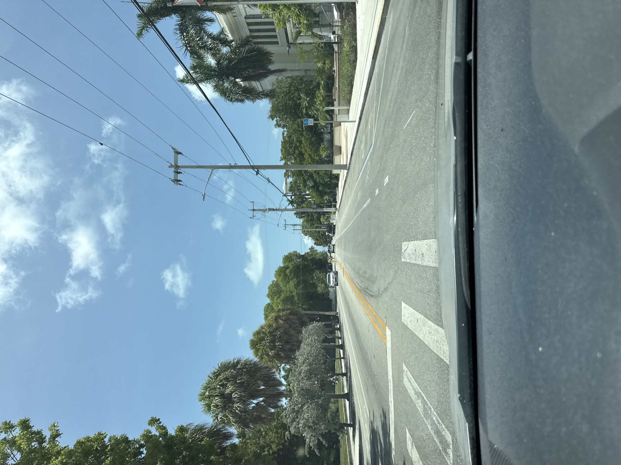 The image shows a view from inside a vehicle driving down a city street, likely in Key West, Florida. The road is paved and has distinct lane markings, including a double yellow line and white pedestrian crosswalk stripes. On the left side of the road, there are lush green trees, palm trees, and a sidewalk. On the right side of the road, there is a modern, light-colored building with columns and an arched entrance. A light blue sign with a wheelchair symbol is visible on the sidewalk. Power lines and utility poles run along the street. The sky is a clear blue with scattered white clouds. Several cars are visible on the road, and a few people appear to be on the sidewalk in the distance. The overall scene depicts a bright, sunny day in a tropical urban environment.