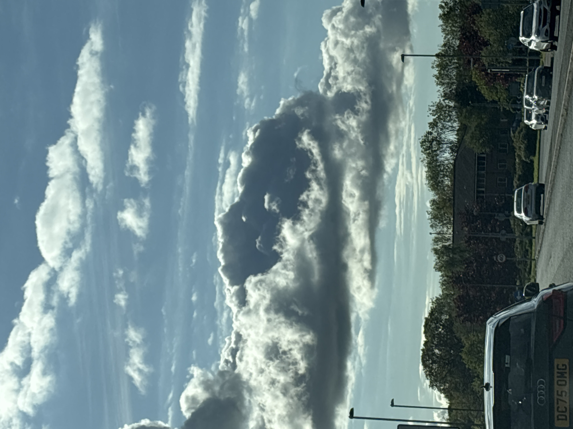 The image shows a daytime sky with prominent cloud formations, photographed from what appears to be a street in Ellesmere Port, United Kingdom. A large, striated cloud formation dominates the center of the frame, ranging in color from bright white to dark gray. Linear cirrus clouds are visible on the left side. The right portion of the image shows street-side trees, a building, streetlights, and several parked vehicles. The lower right corner of the frame contains the rear of a dark-colored car, including its license plate "DC75 OMG". The overall impression is of a partly cloudy sky observed from an urban or suburban roadside.