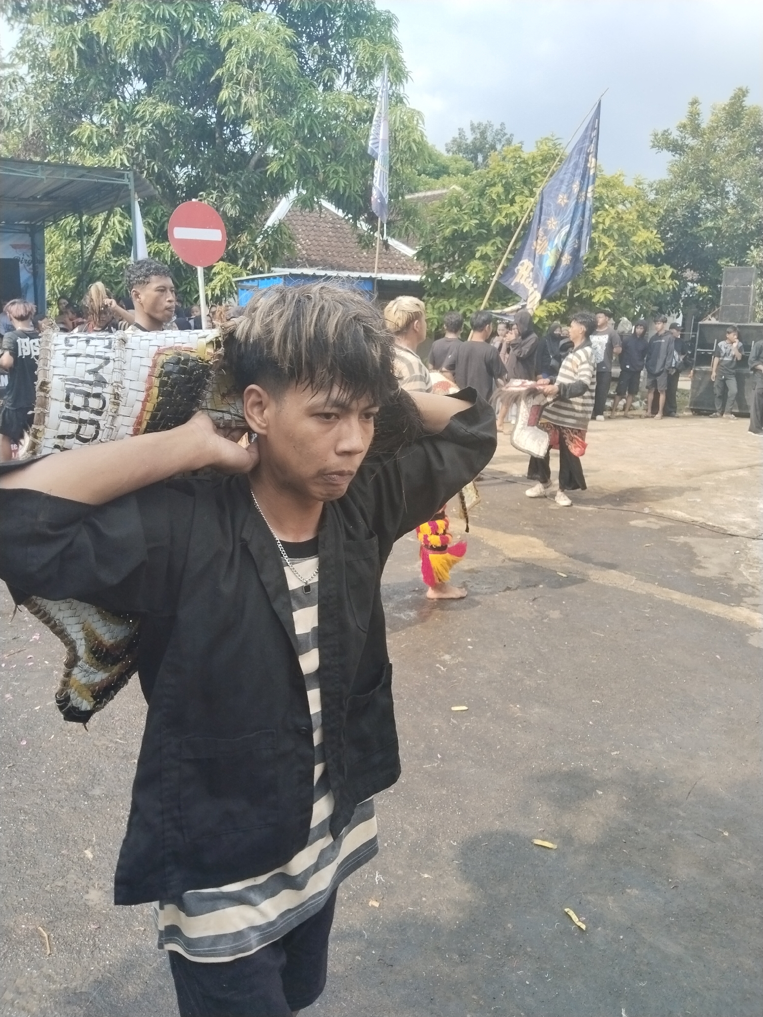 The scene is in Pendem, Indonesia, with a group of people gathered on a paved area or road. The focus is on a young individual with distinctive two-toned hair, wearing a striped shirt, a black jacket, and carrying a large, rectangular woven object on their shoulders. In the background, there are several other people, some wearing similar attire, a flag, and a building. A red and white no-entry sign is visible in the background. There is debris, seemingly spent firecrackers, scattered on the ground. The overall impression is of a cultural event or celebration.