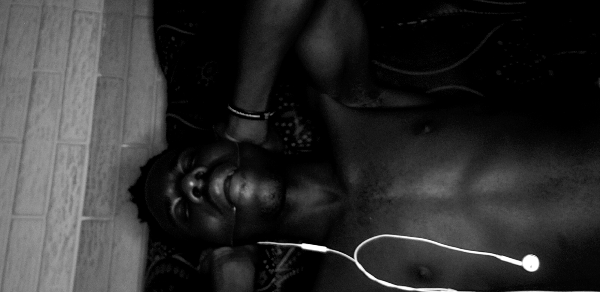A shirtless man is lying on his back with a pair of headphones plugged in. He is wearing a bracelet on his left wrist. The location may be Narayi, Nigeria, given the prompt's specification. The man is lying on a patterned surface next to a tiled wall.