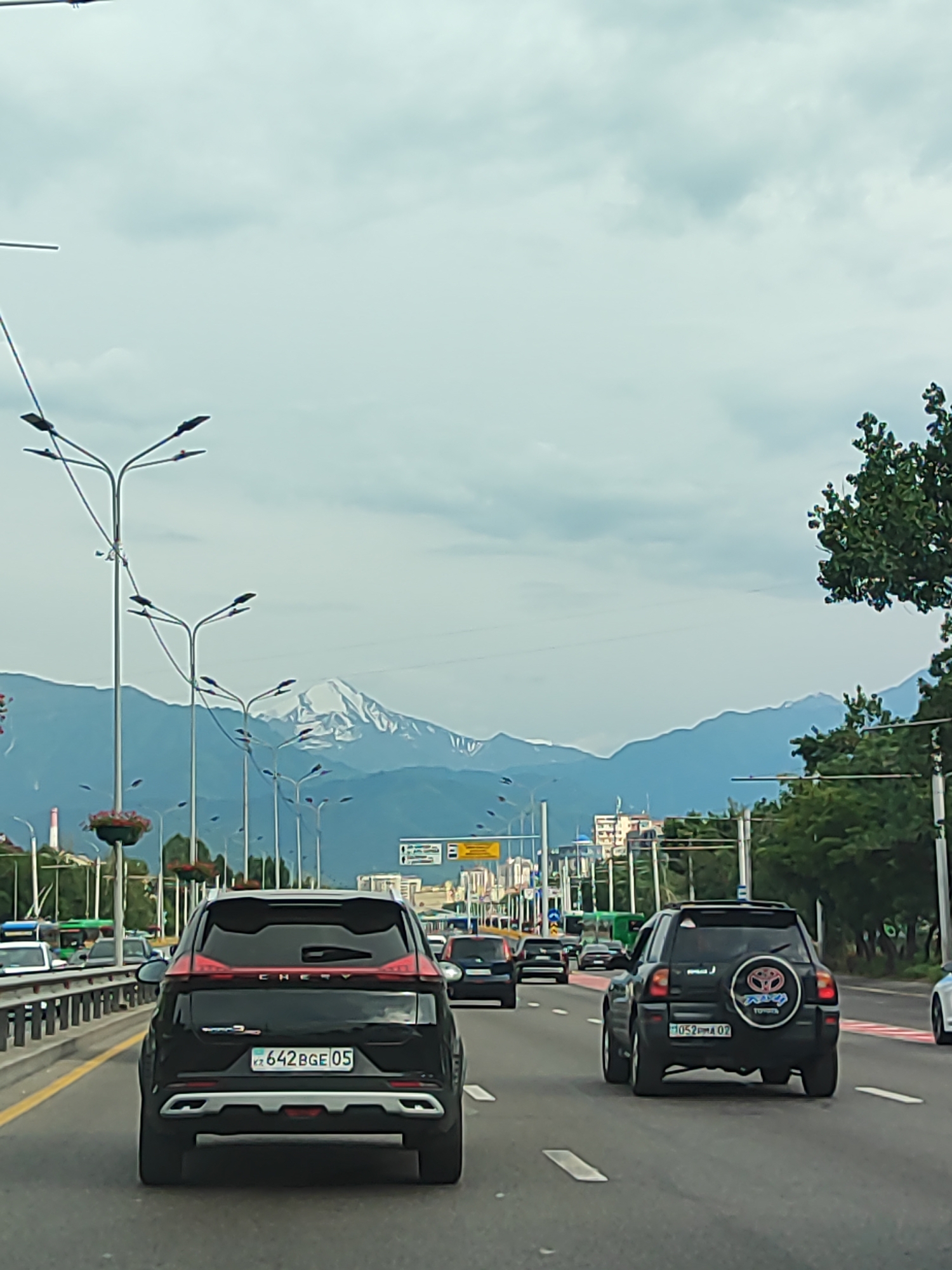 This is a street-level, rear-facing view of traffic on a multi-lane road, likely in Almaty, Kazakhstan. The sky is overcast, suggesting a cloudy day. In the background, majestic snow-capped mountains are visible, providing a dramatic natural backdrop to the urban scene.
The primary subjects are several cars on the road. Closest to the camera is a black Chery SUV with a distinctive red light bar across the rear. Its license plate reads "KZ 642 BGE 05". To its right, a black Toyota RAV4 is visible, featuring a spare tire cover with the Toyota logo and "RAV4" written on it. Its license plate is "KZ 052 RMA 02". Other vehicles are further down the road, indicating moderate traffic flow.
The setting is an urban environment with a highway infrastructure. Tall streetlights line the road, and there are some buildings and trees visible on the sides. The road has clear lane markings. The overall impression is of a typical day in a city that borders impressive mountain ranges. There are no people clearly visible in the image, and the focus is on the vehicles and the landscape.