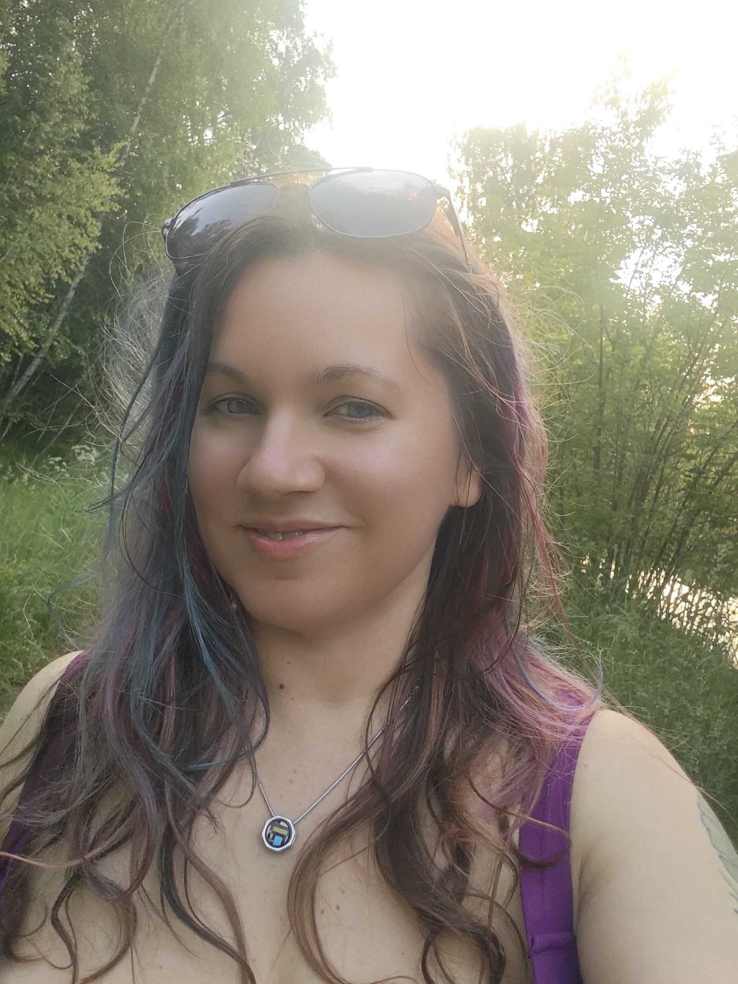 A woman is taking a selfie outdoors in what appears to be a natural setting with trees and foliage. She is wearing a purple tank top and a silver necklace with a pendant. Sunglasses are resting on top of her head. Her hair is long and wavy with streaks of purple and blue. The background is blurred, suggesting she is in motion or the focus is on her. The sun is visible in the upper right corner of the image, creating a bright glare. The exact location within Водник Снт, Russia, cannot be determined from the visual information provided.