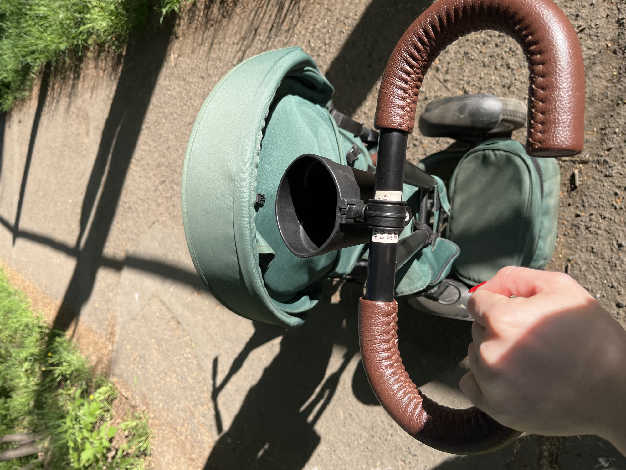 The image shows a top-down view of a stroller or tricycle-like object on a paved surface, likely in an urban area, suggested by the presence of grass and a sidewalk. The stroller features a green canopy and seat, and a brown, leather-wrapped handlebar. A black cup holder is attached to the frame. A person's hand is visible, positioned near the handlebar. The location is likely the city of Chaikovsky, Russia based on inscriptions in Cyrillic present on the stroller’s frame. The scene appears to be taken outdoors on a sunny day, casting shadows across the pavement.
